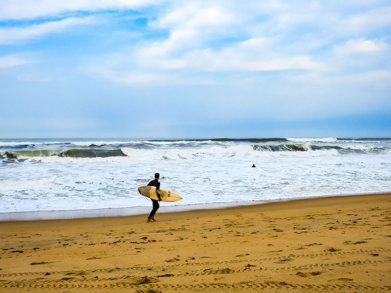 Surfer carrying yellow board walks across sandy beach toward breaking ocean waves.