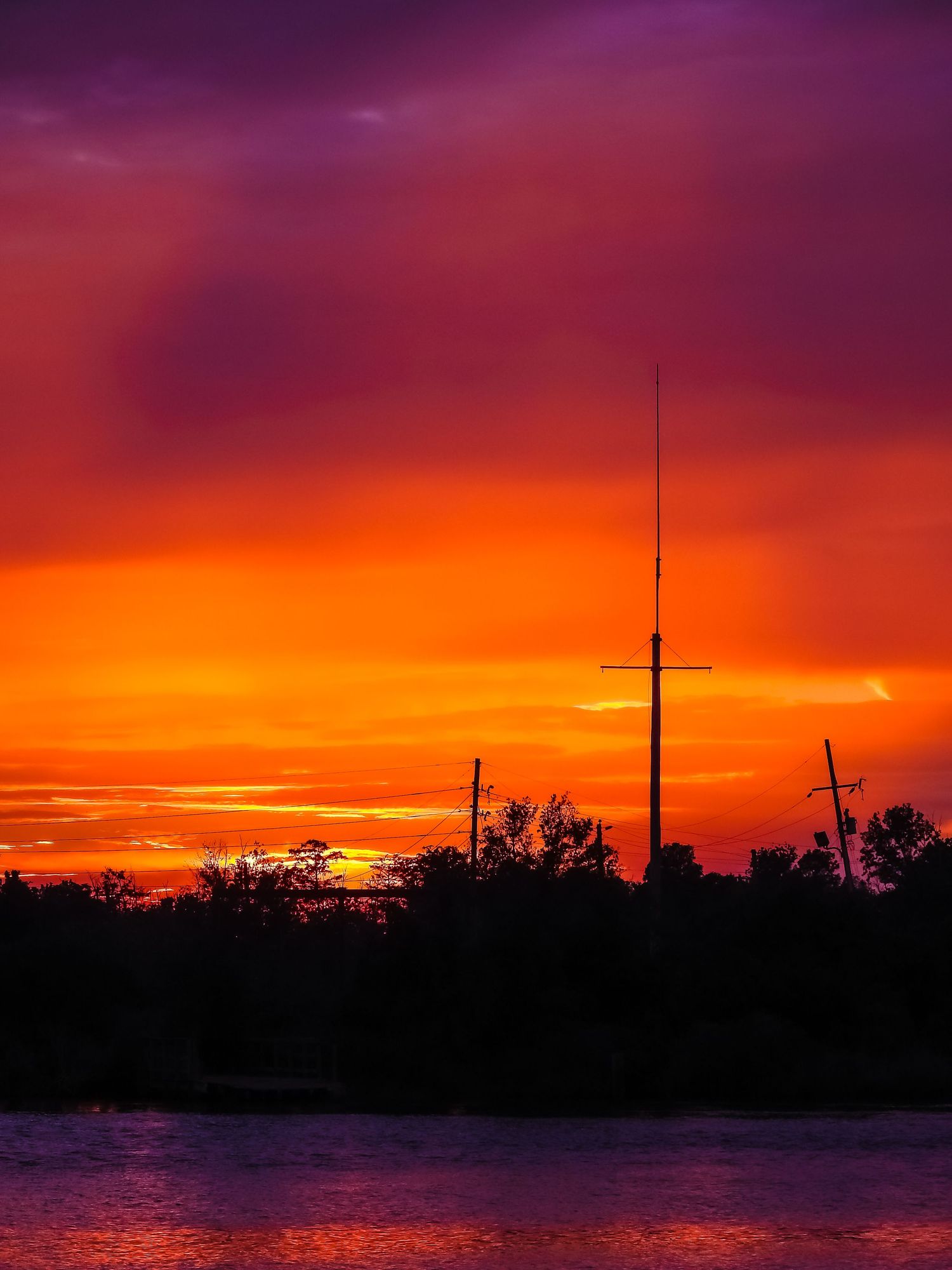 Dramatic orange and purple sunset silhouettes trees and a radio tower against the water.