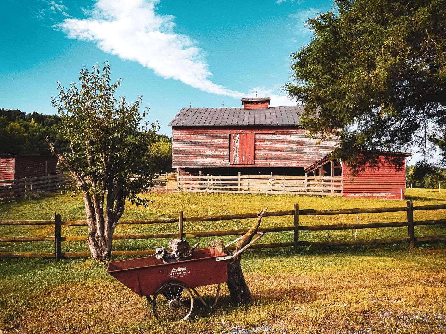 Red farmhouse with wooden fence and vintage wheelbarrow in rustic countryside setting.