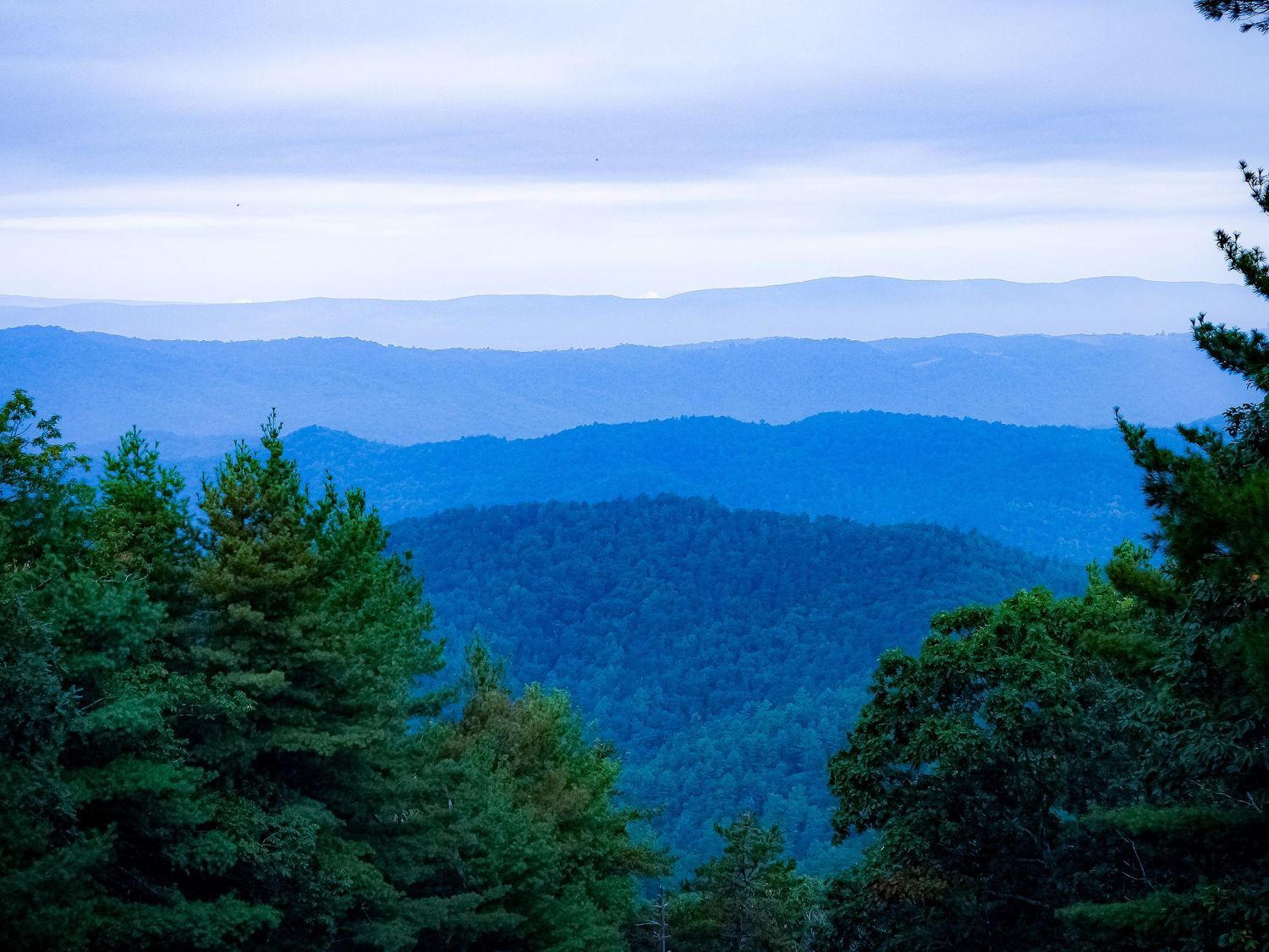 Misty blue mountain ranges create a layered effect against cloudy sky and evergreen trees.