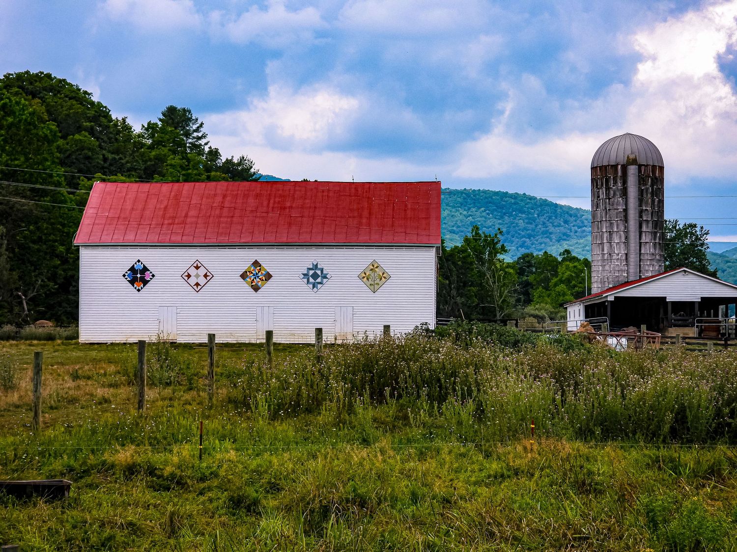 White barn with decorative windows stands beside tall silo against mountain backdrop.