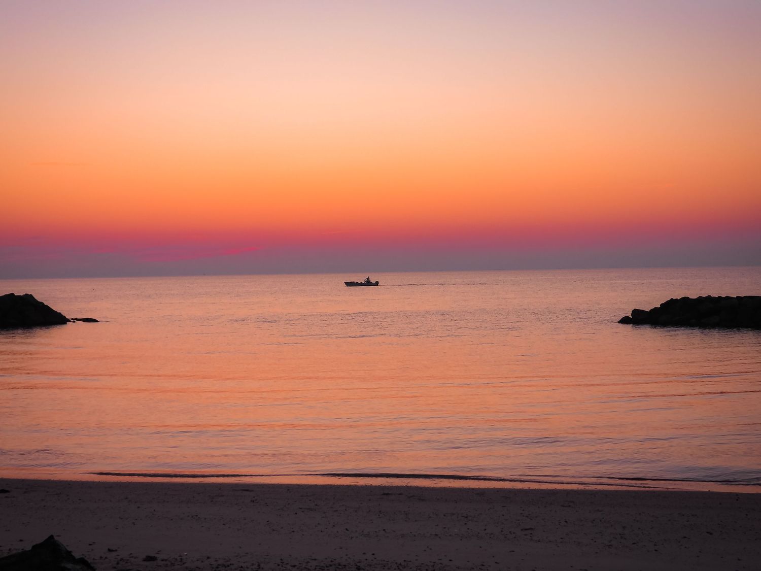 Silhouette of a boat on horizon between rocky outcrops during a pastel orange sunset.
