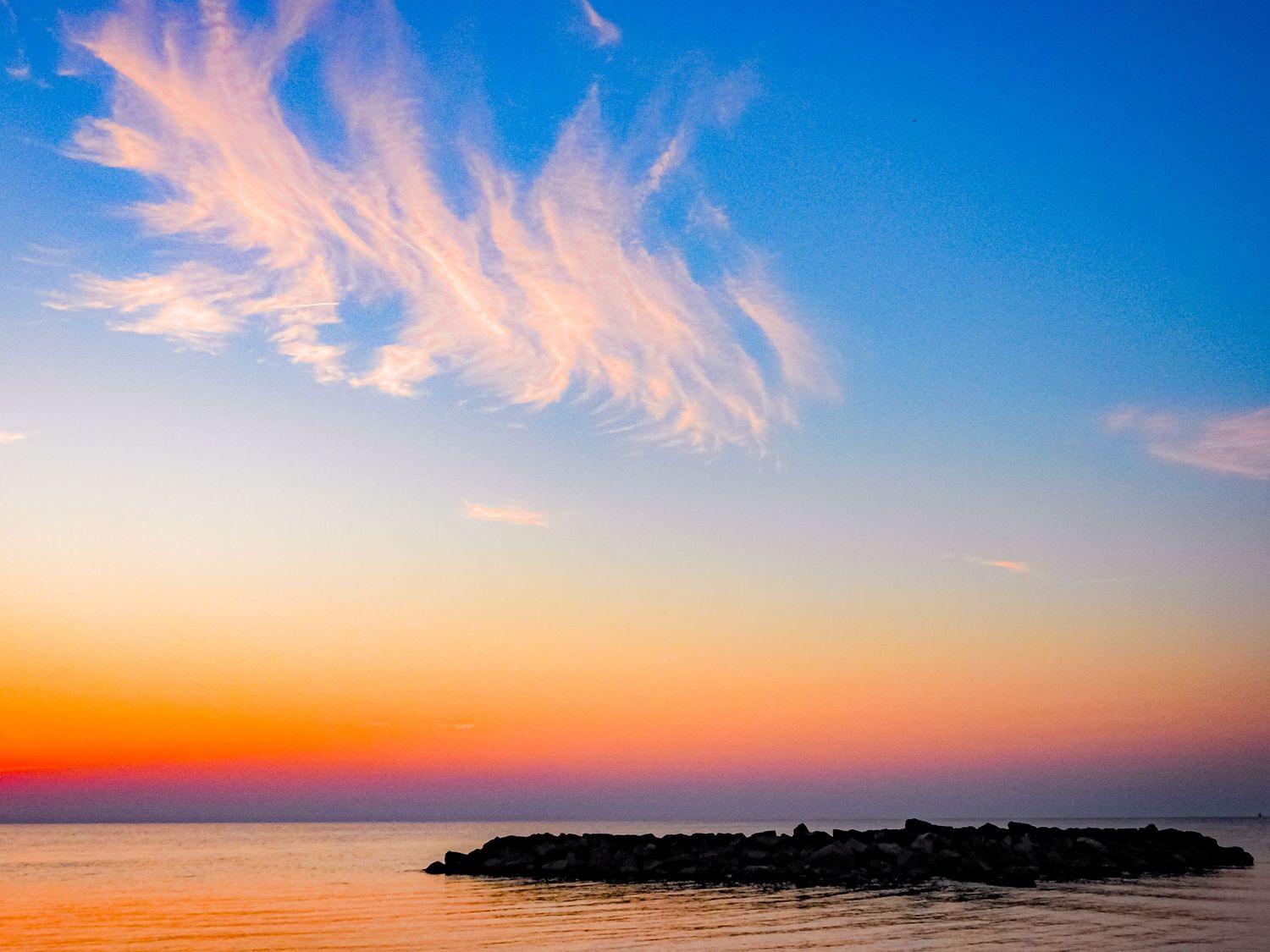 Dramatic pink clouds streak across blue sky over dark ocean jetty at sunset.