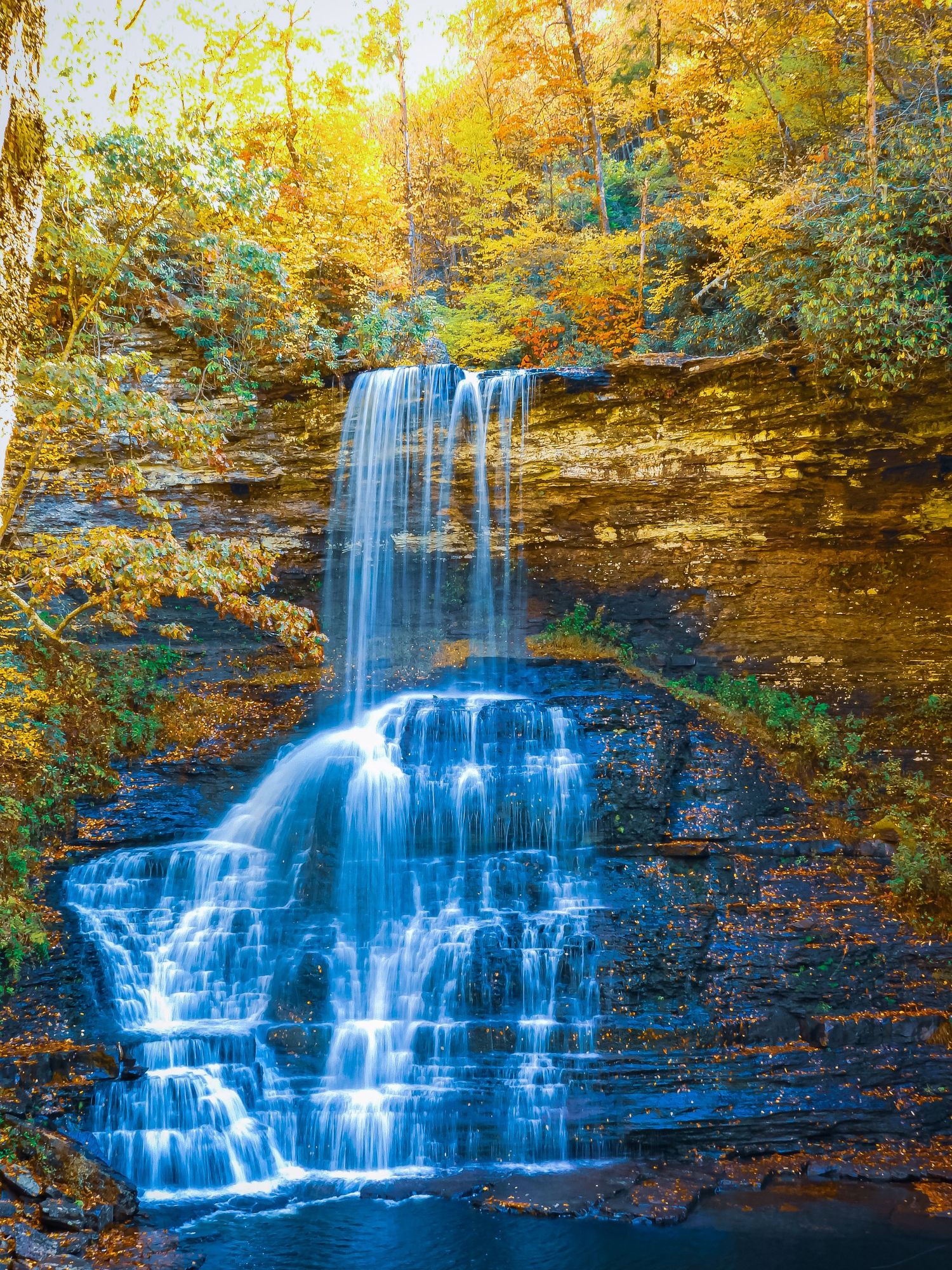 Multi-tiered waterfall cascades over rocky cliffs surrounded by vibrant autumn foliage.