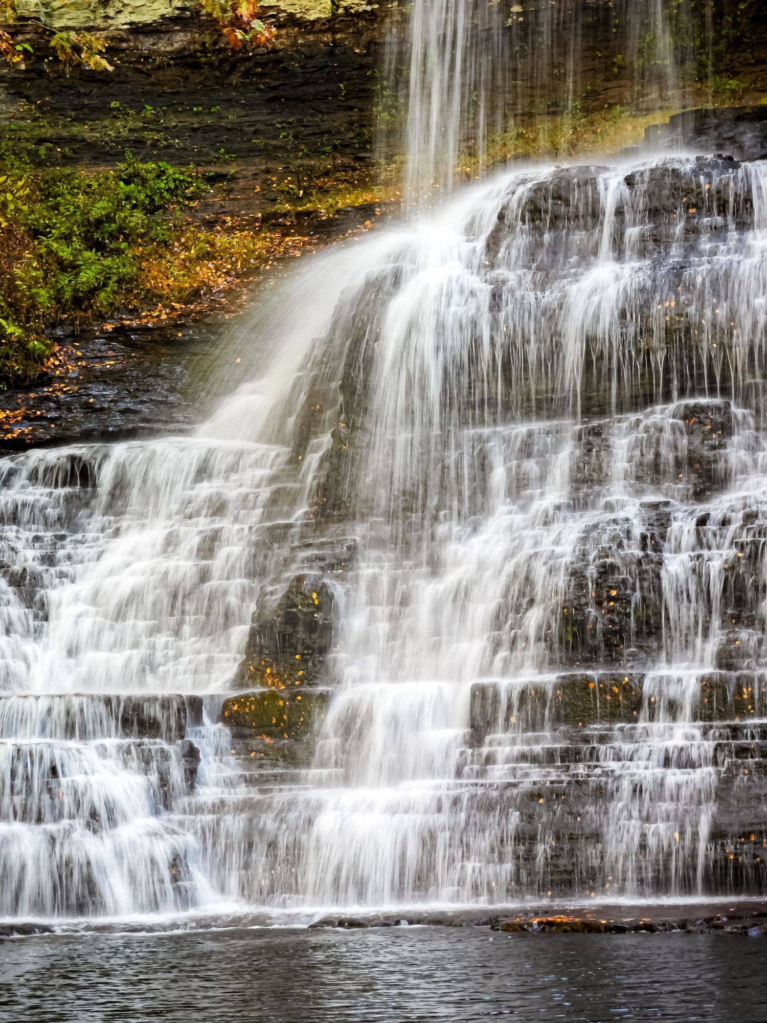 Cascading waterfall flows down rocky ledges with multiple tiers of white rushing water.