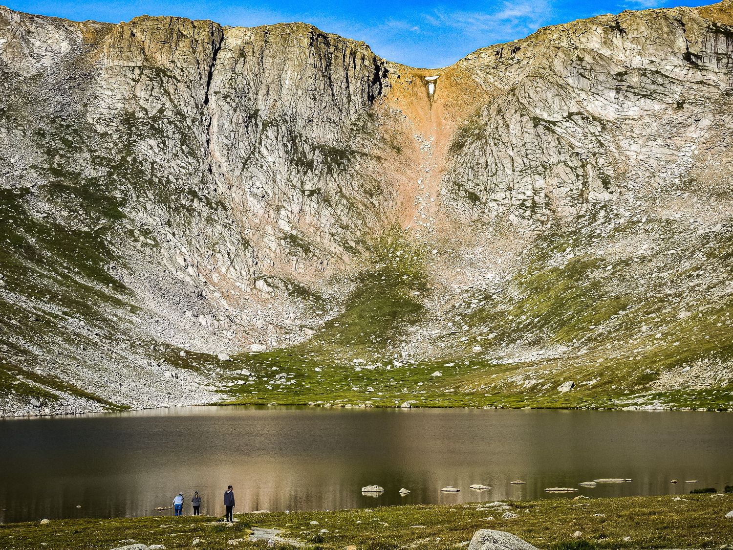 Steep rocky cliffs surround an alpine lake with hikers providing scale at the water's edge.