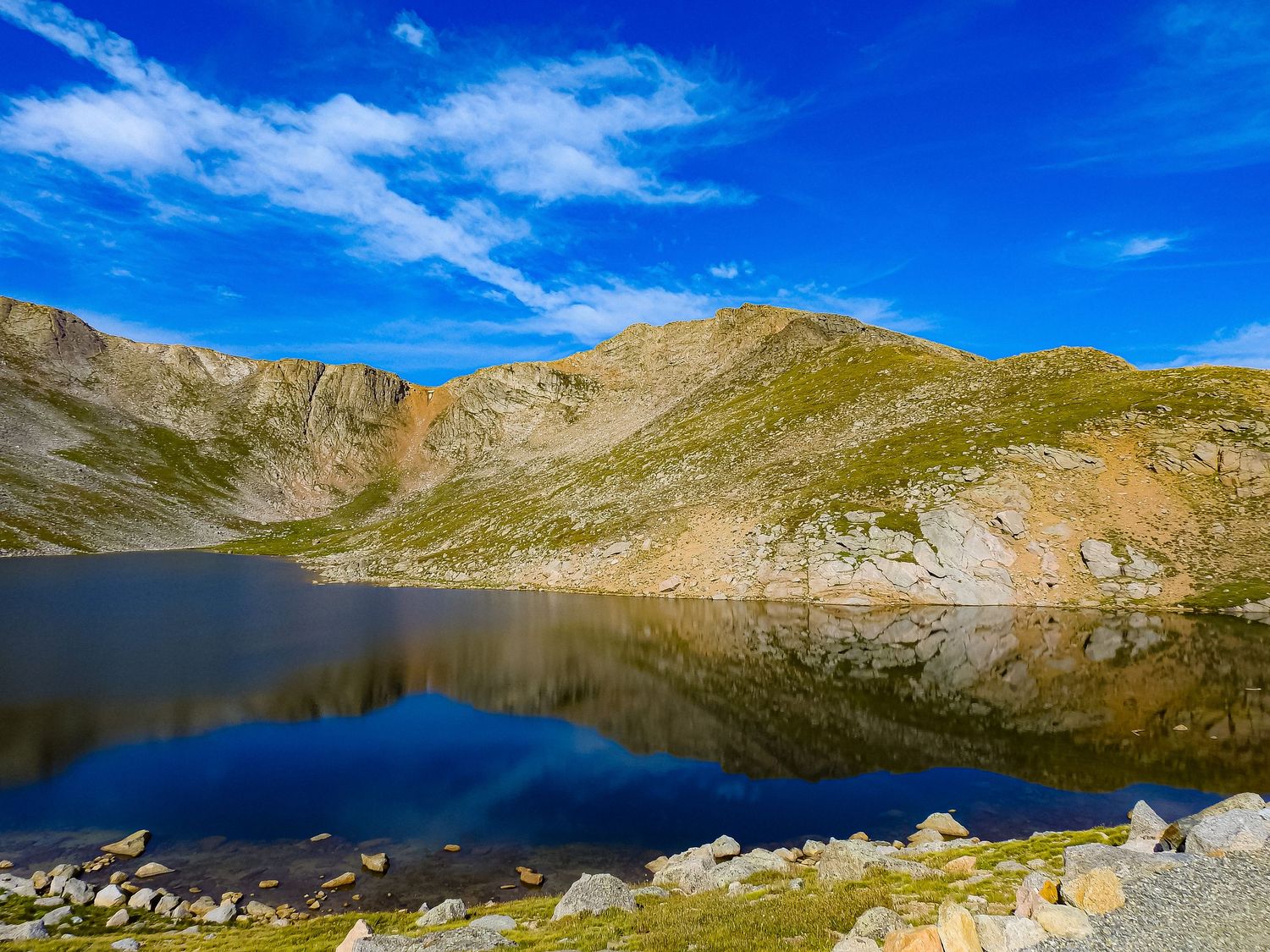 Crystal clear mountain lake mirrors surrounding peaks and bright blue sky in alpine setting.