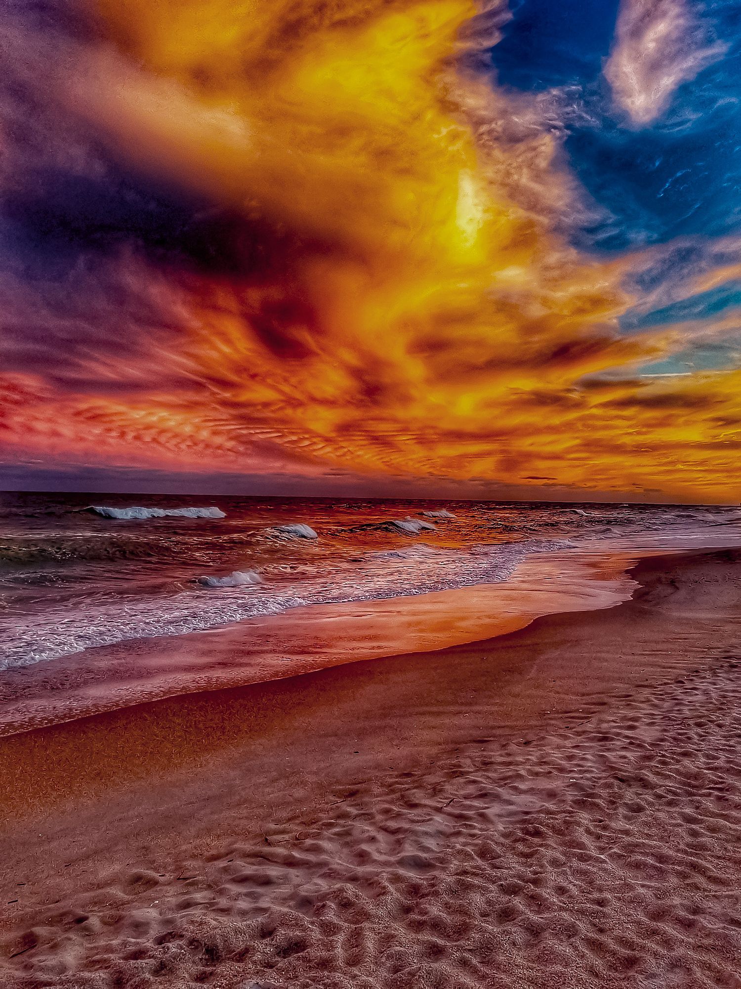 Vibrant red and orange sunset sky reflects on wet beach sand as waves roll in.