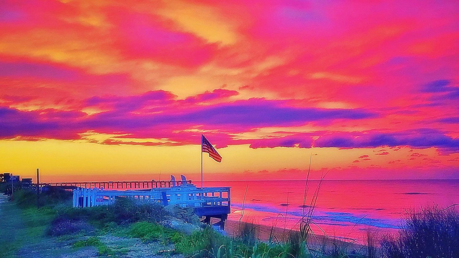 Vibrant pink and purple sunset sky over a coastal pier with an American flag silhouetted against dramatic clouds.