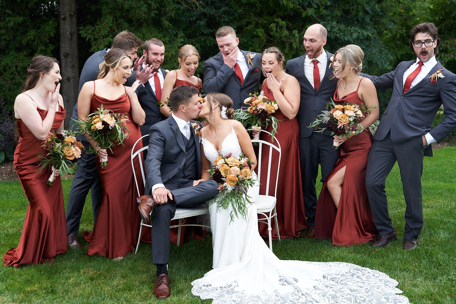 Wedding party in burgundy dresses and dark suits gathered outside for formal group photo. Wedding party cheers while bride and groom kiss.
