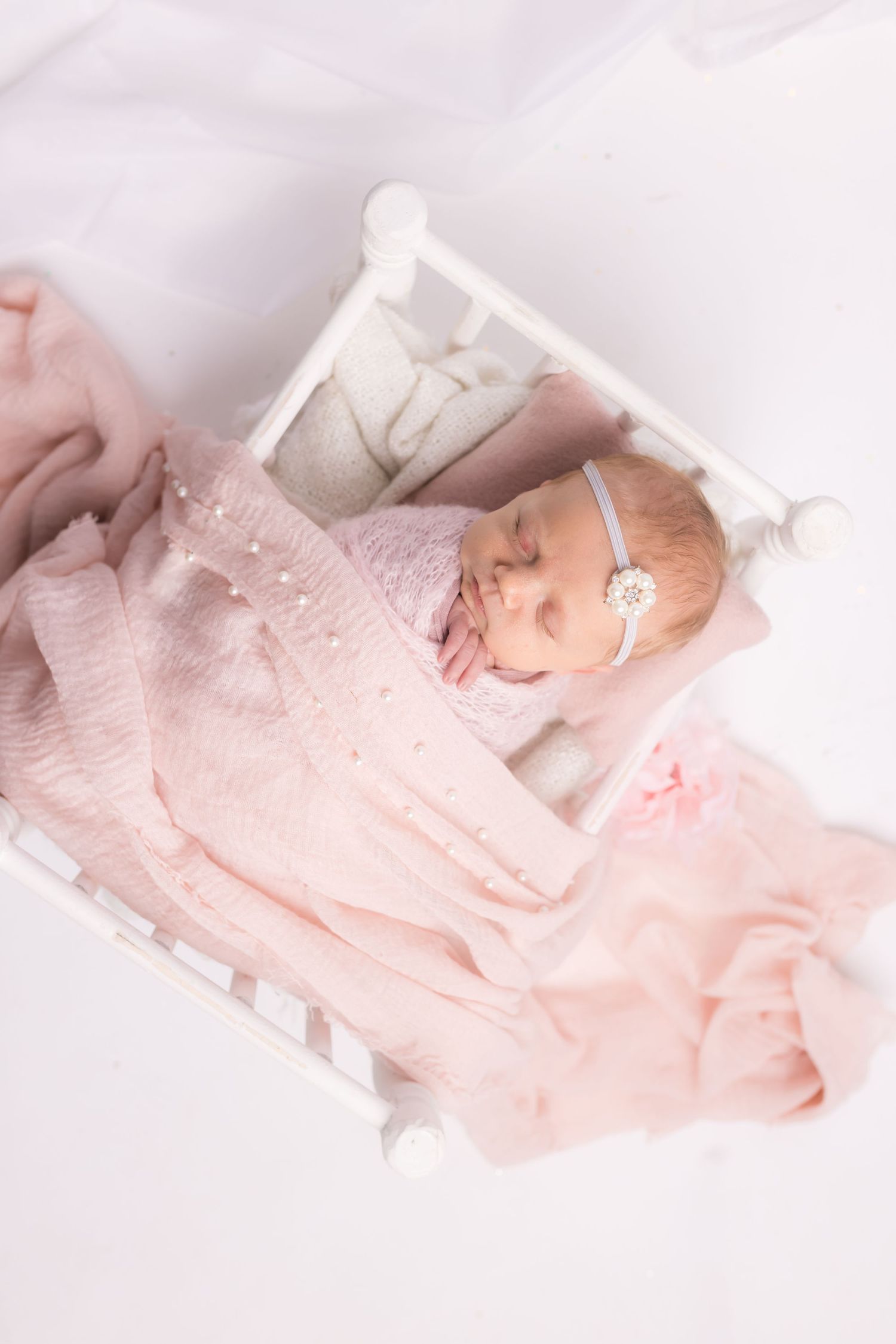 Sleeping newborn in pink outfit lying in miniature white bed with delicate accessories.