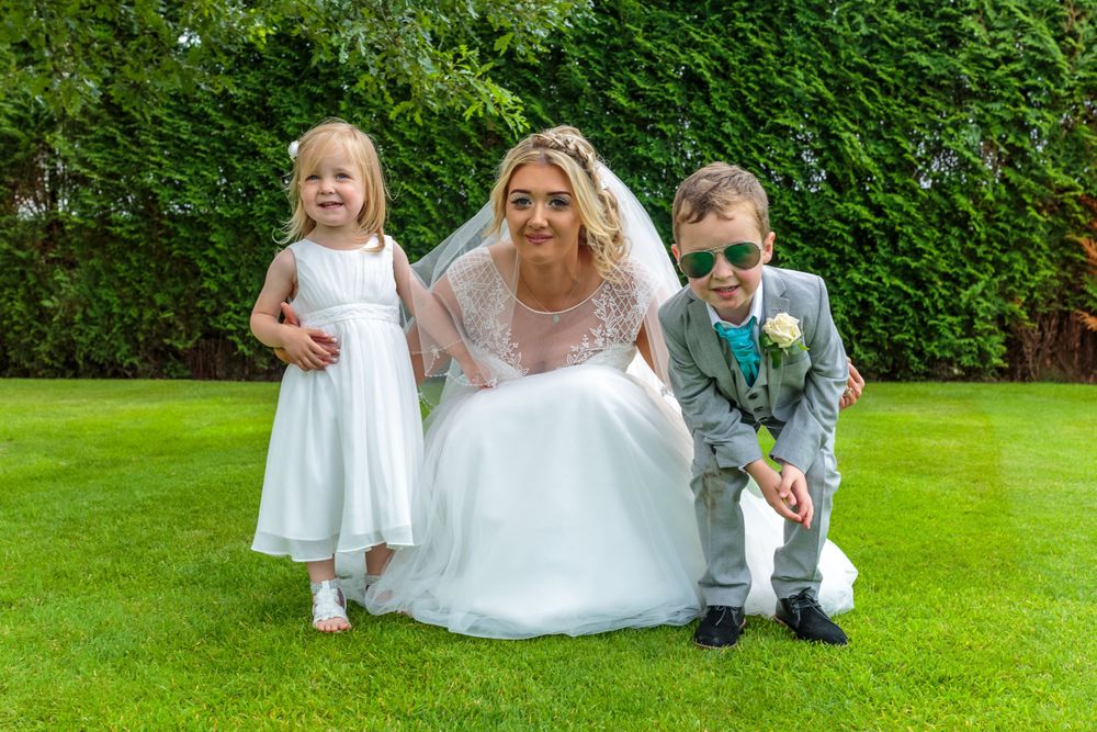 bride crouches down for a photograph with the little flower girl and the page boy wearing mirrored sunglasses