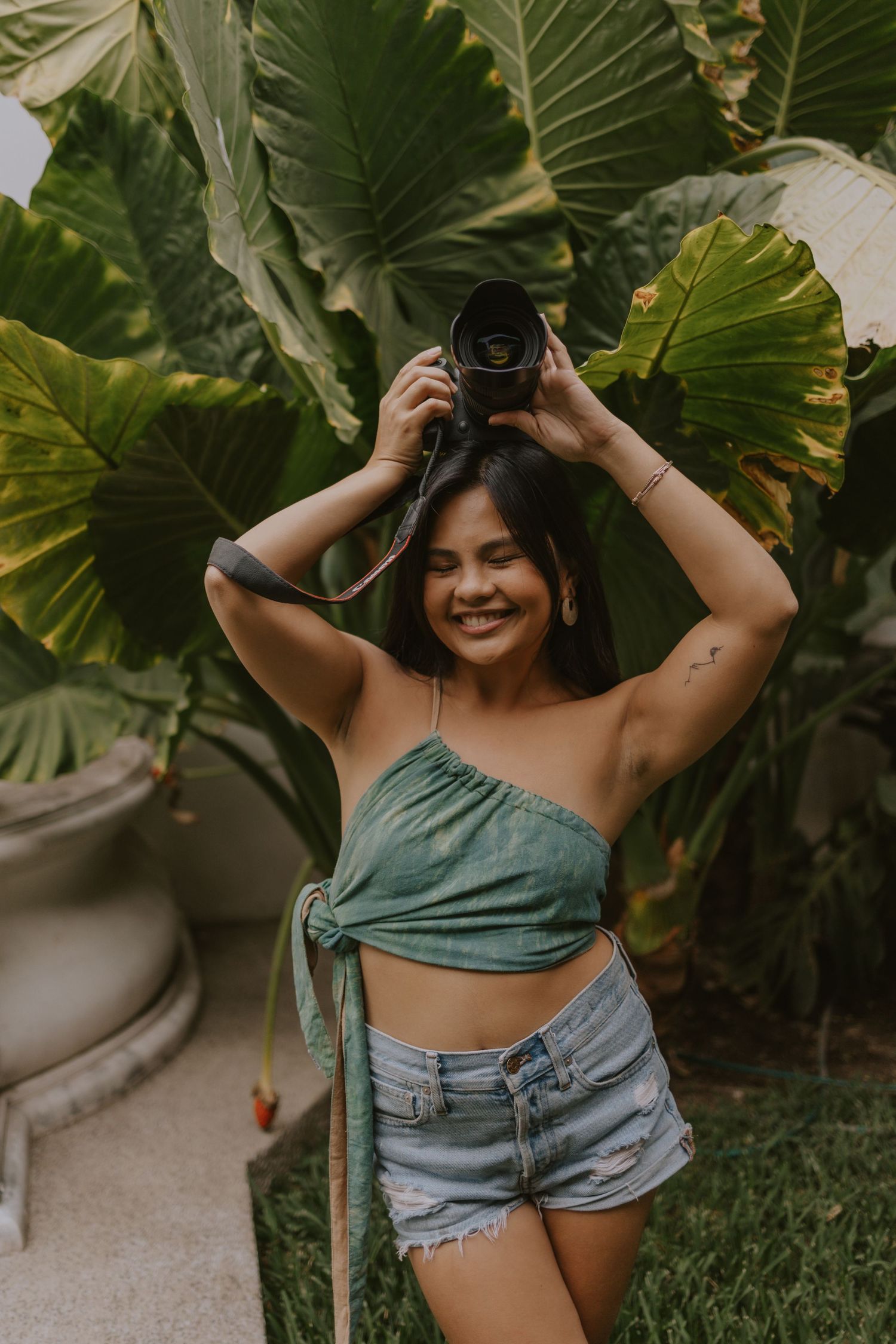 A photographer in casual summer attire poses with a camera against tropical foliage.