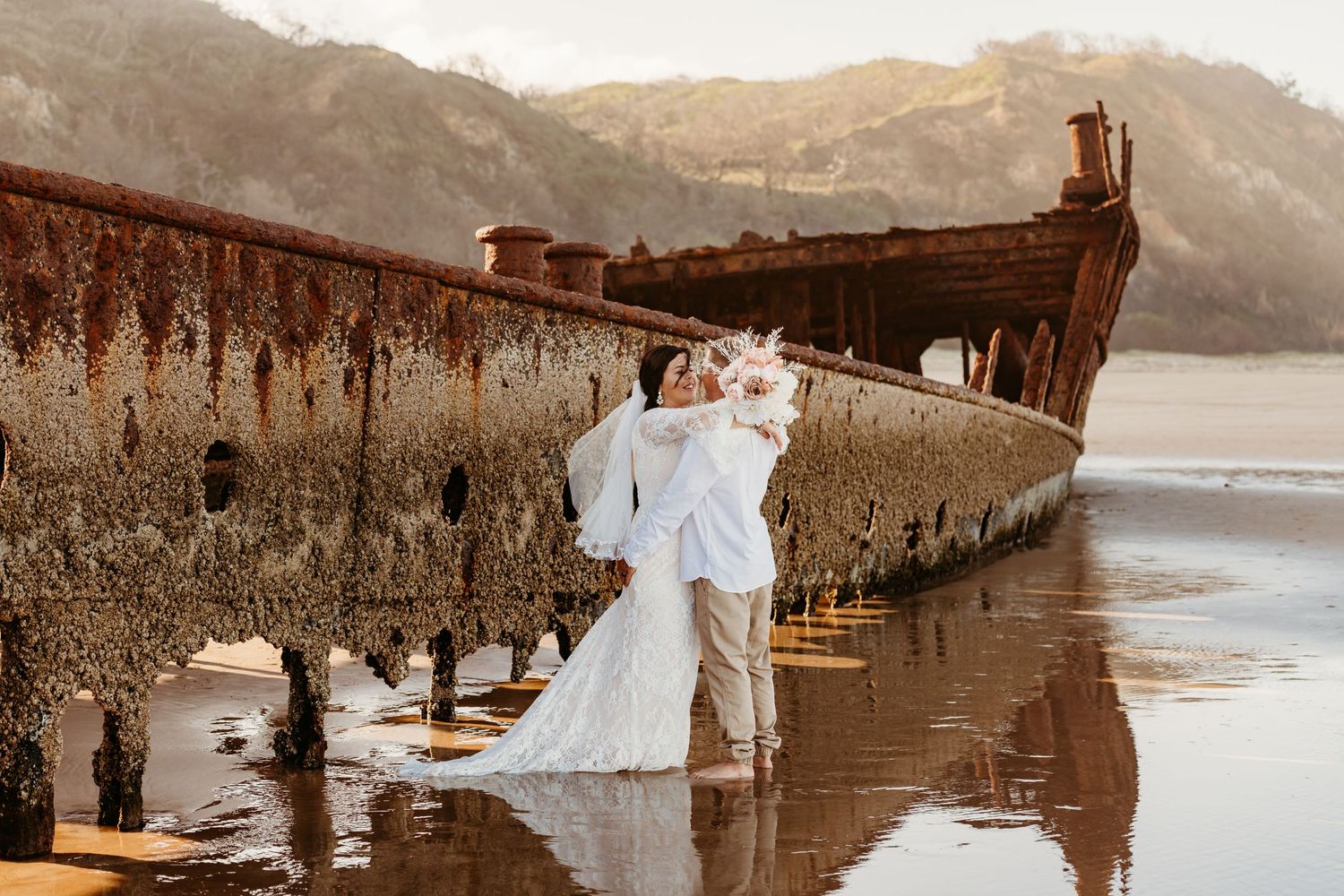Wedding couple standing by rustic shipwreck on beach during golden hour.