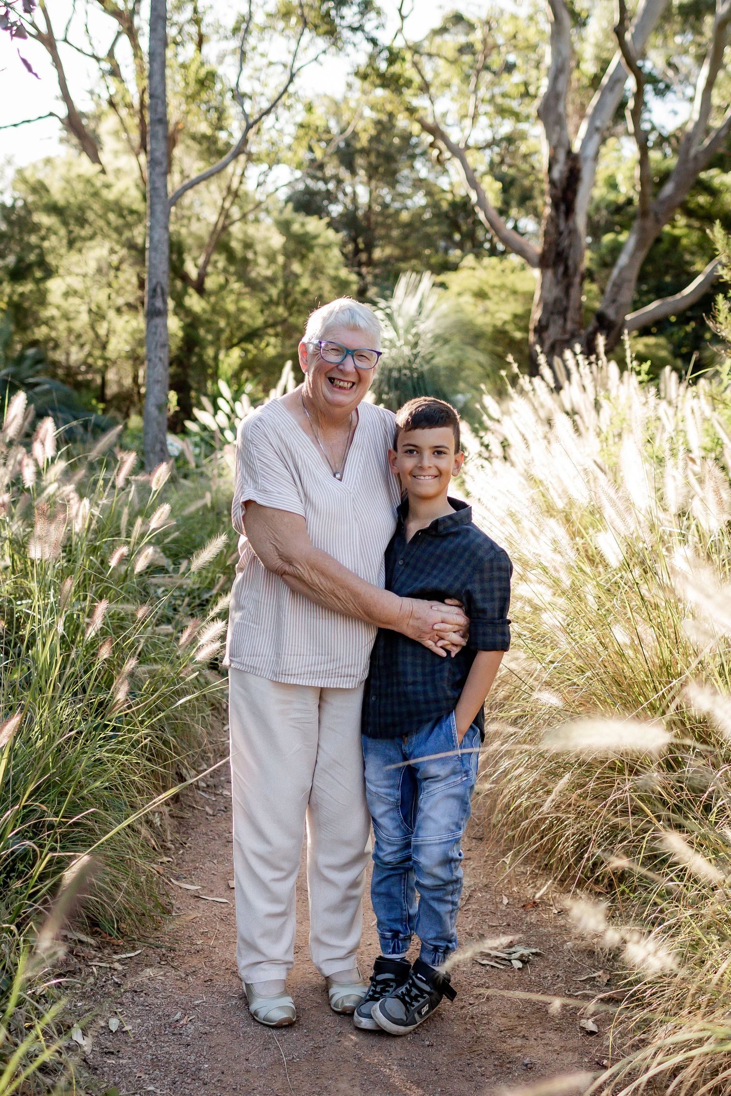Two people standing together on a dirt path with sunlit pampas grass and trees in the background.