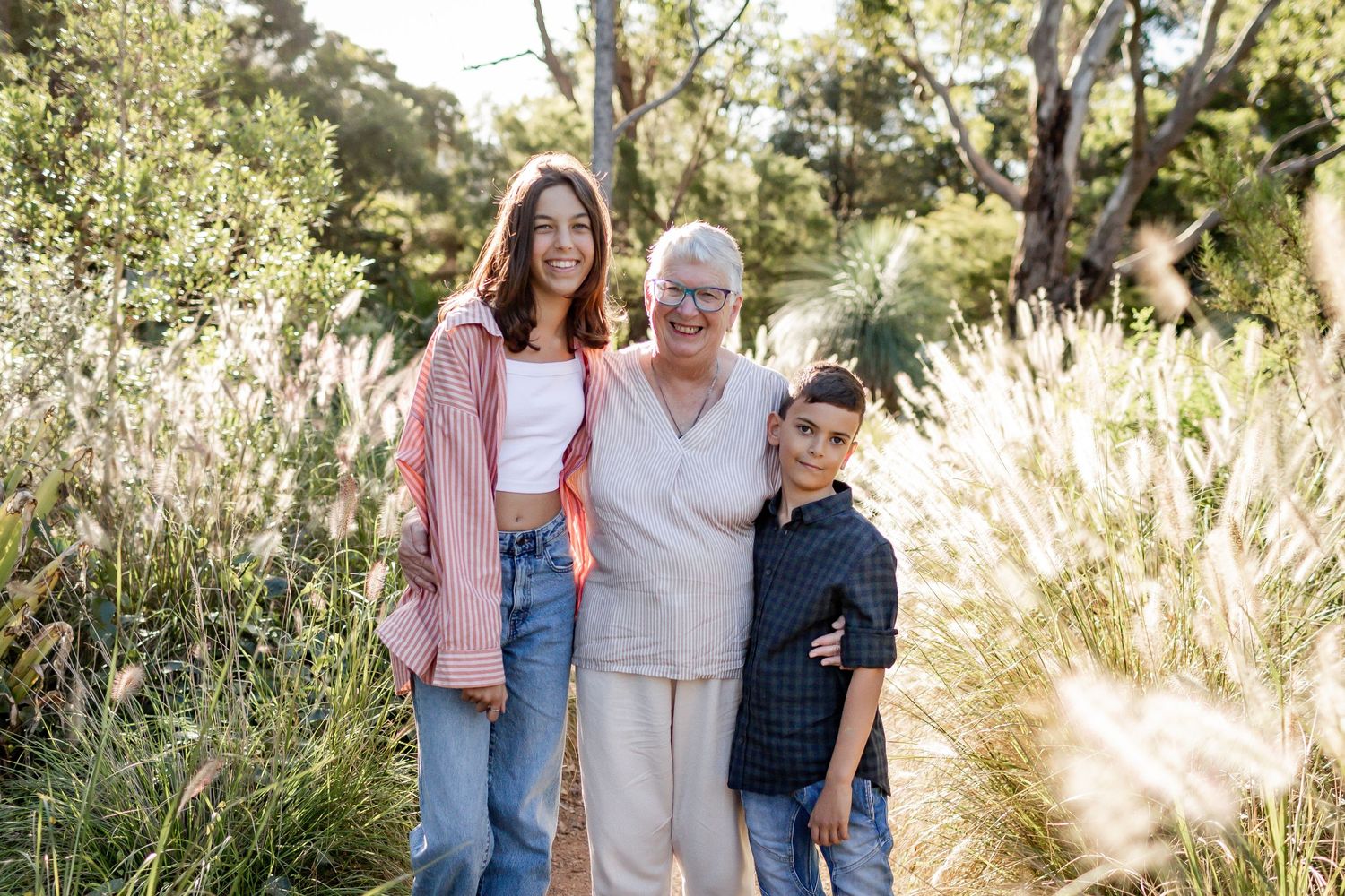 Three people pose together on a sunlit nature path with grass and trees in the background.