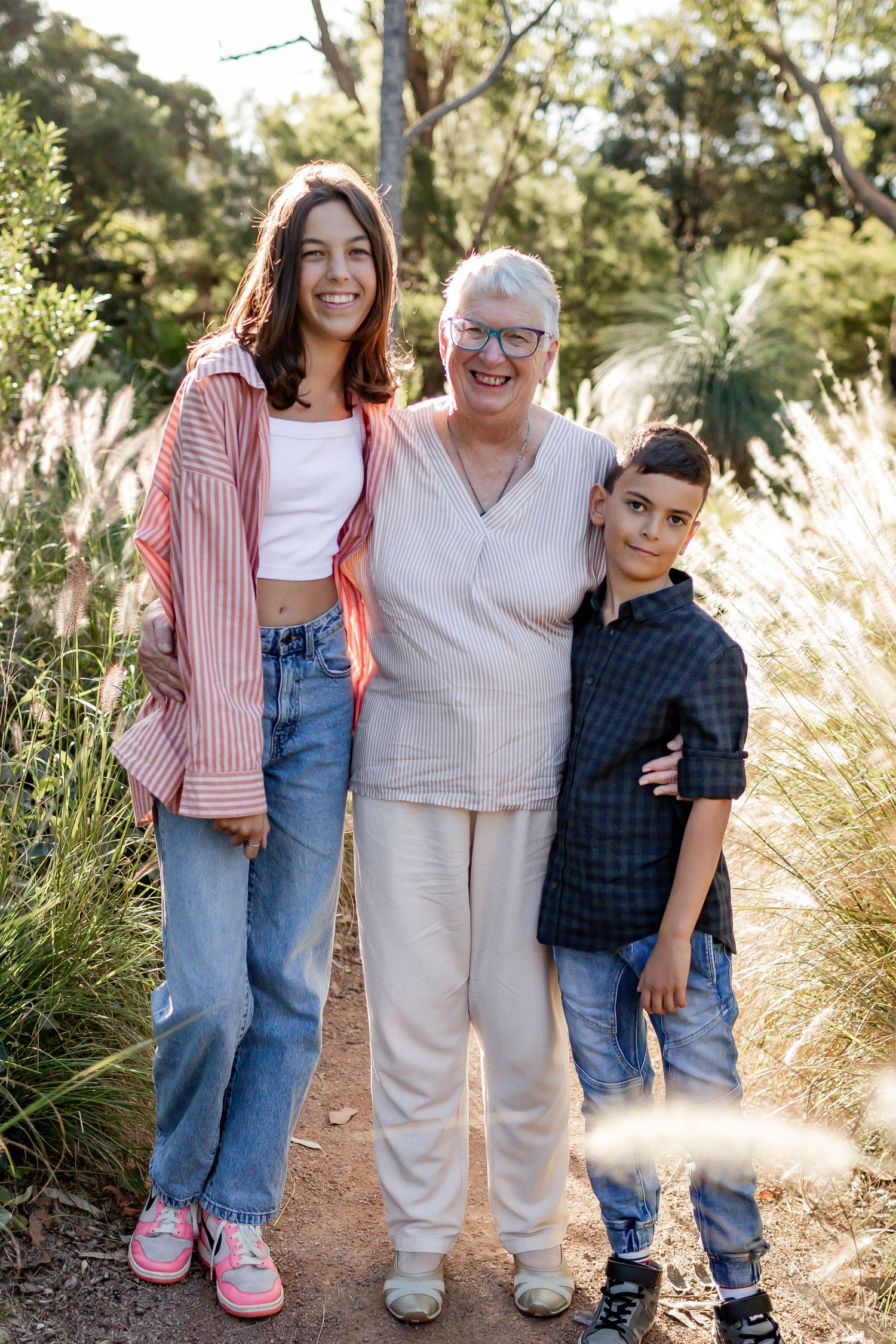 Three generations pose together on a sun-dappled nature trail with golden grass.