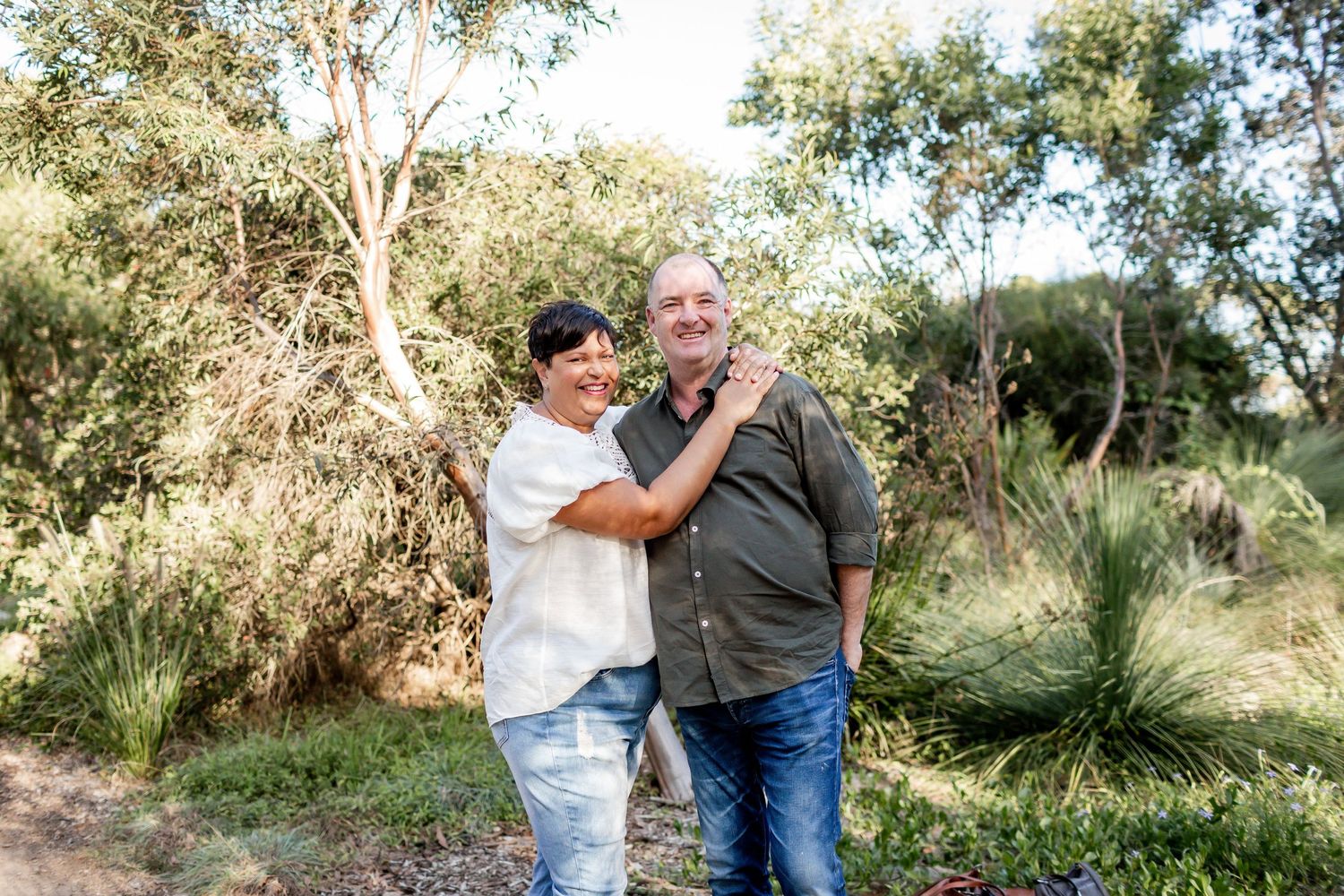 Two people embrace while standing on a sunny nature path surrounded by trees.