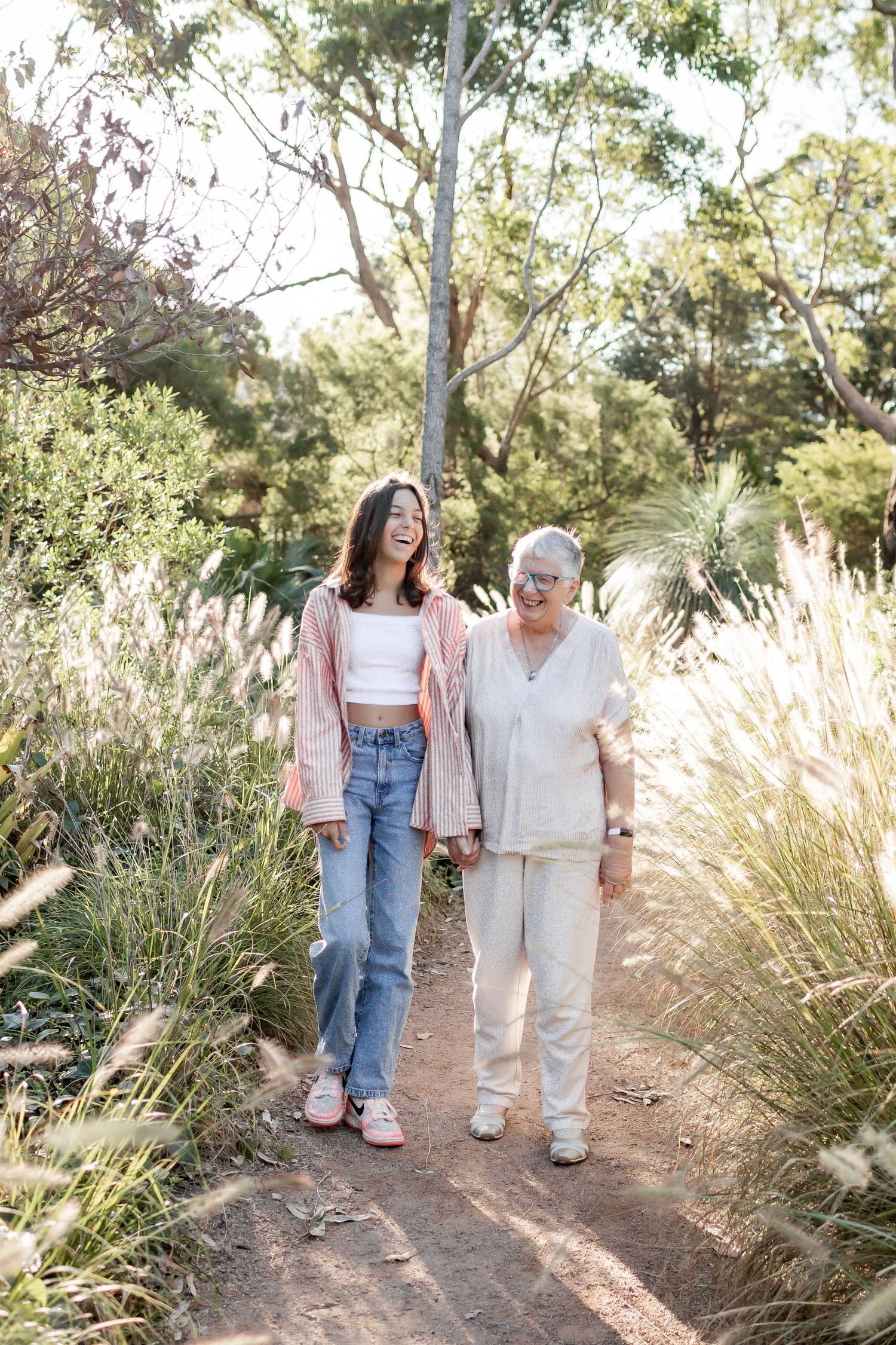 Two people walking together on a nature trail surrounded by tall ornamental grasses and eucalyptus trees.