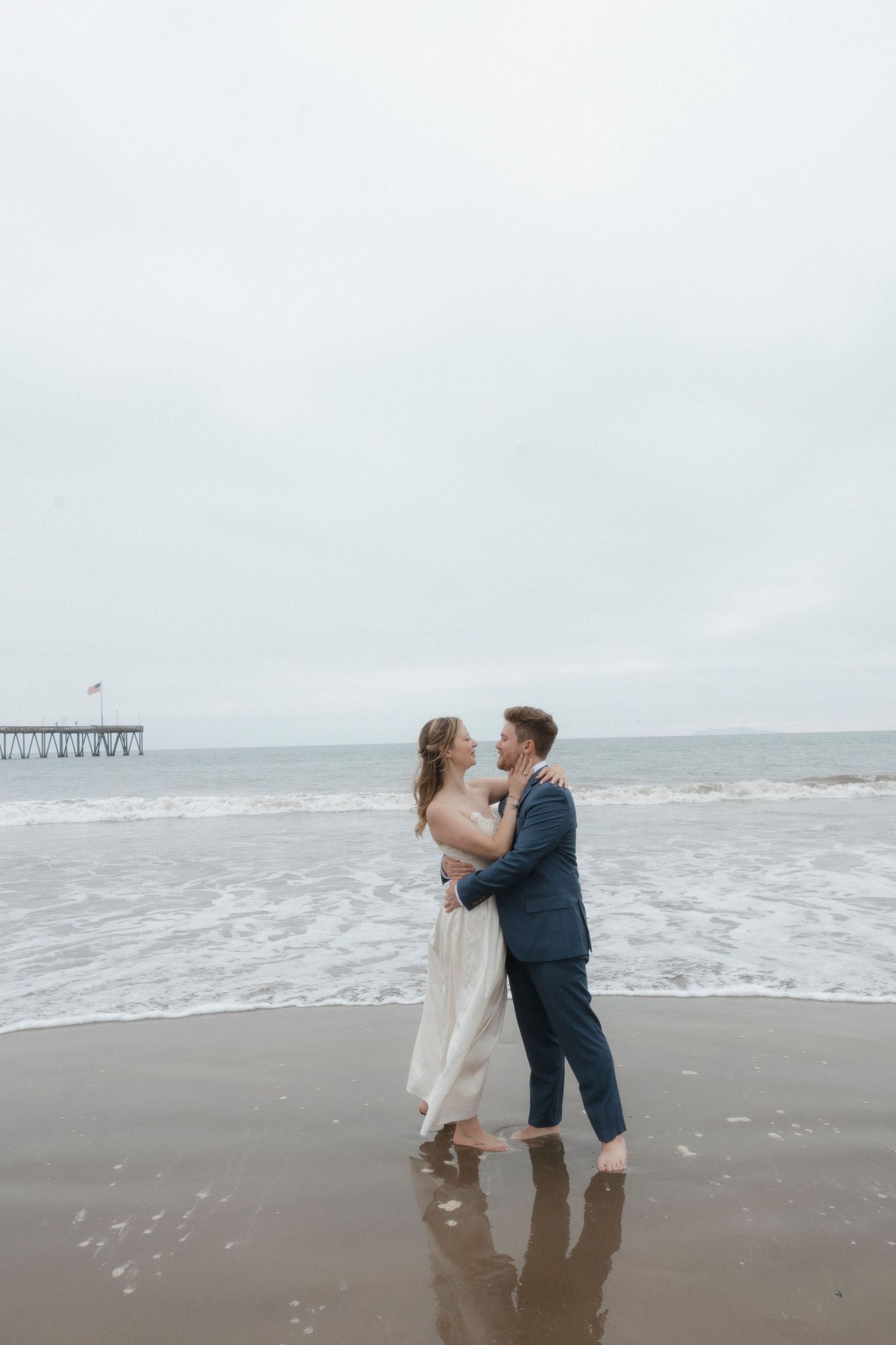 A couple embraces on a cloudy beach with waves and a pier in the background.
