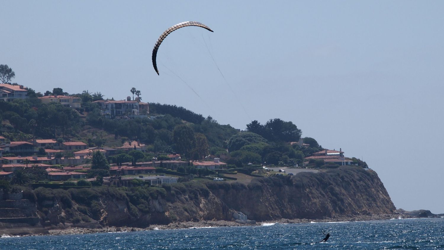Paraglider soaring over a coastal cliff with ocean views and hillside homes on a clear day.