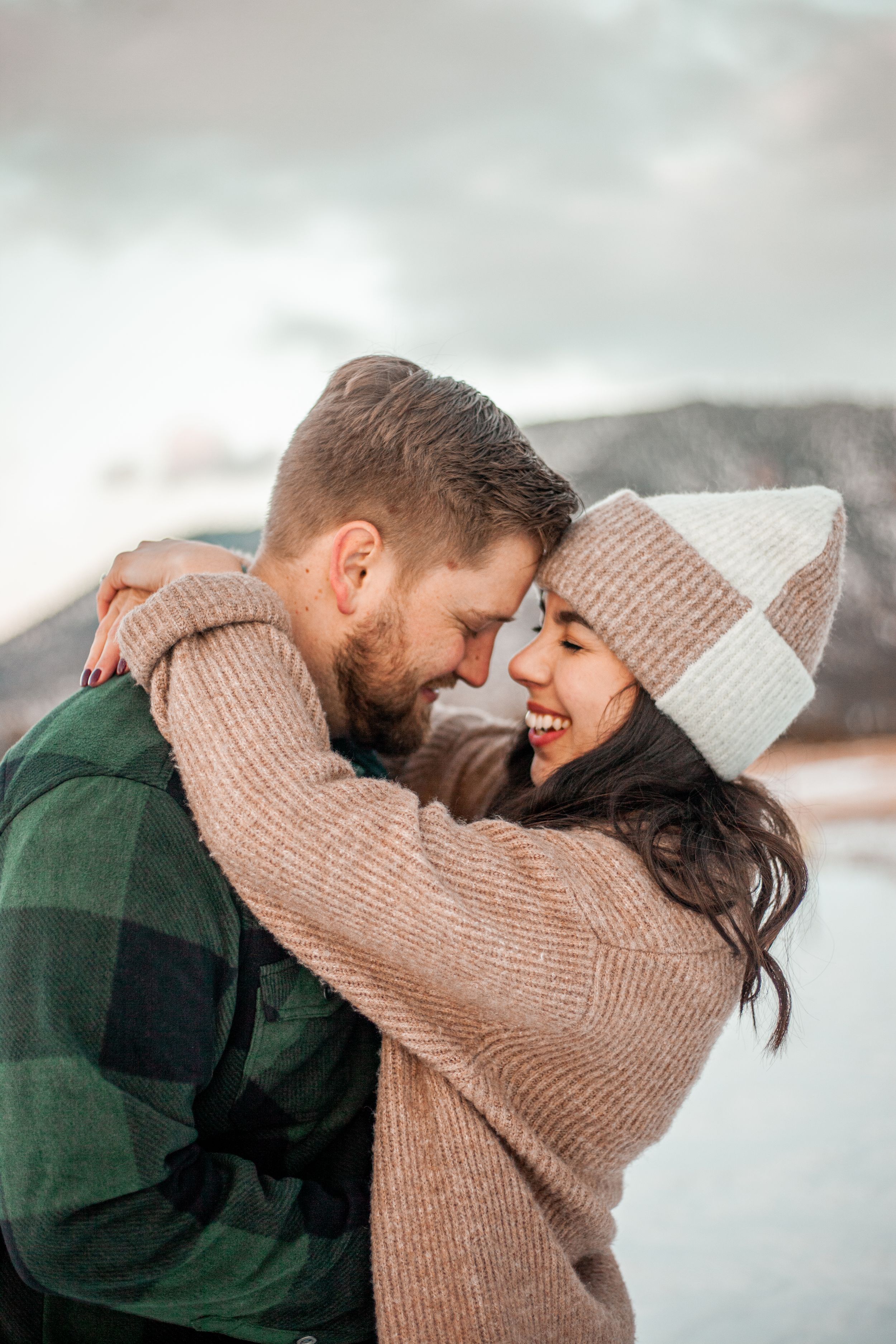 Cozy winter embrace between couple wearing knit sweaters and beanies against snowy backdrop.