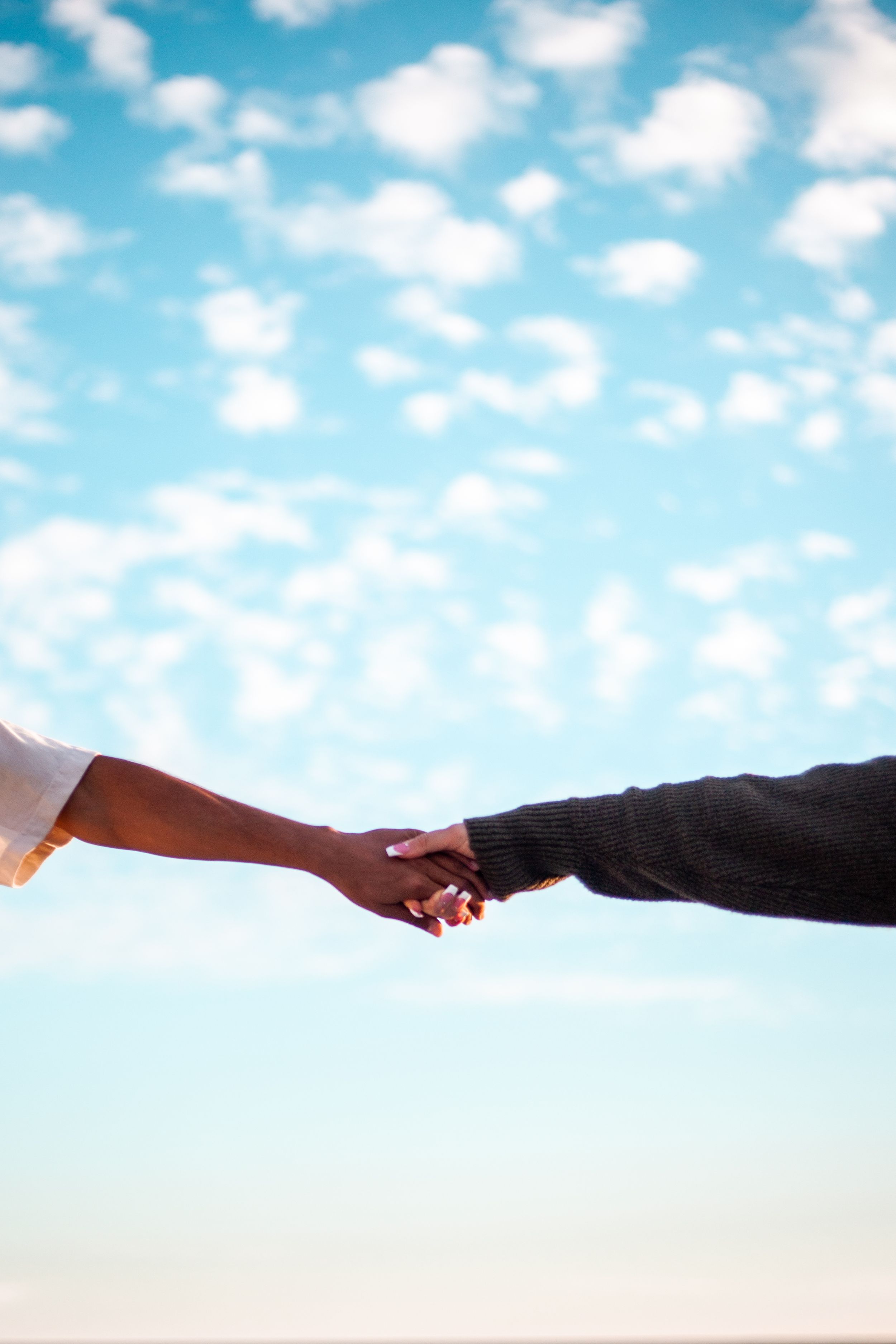 Two hands reaching towards each other against a bright blue sky with fluffy clouds.