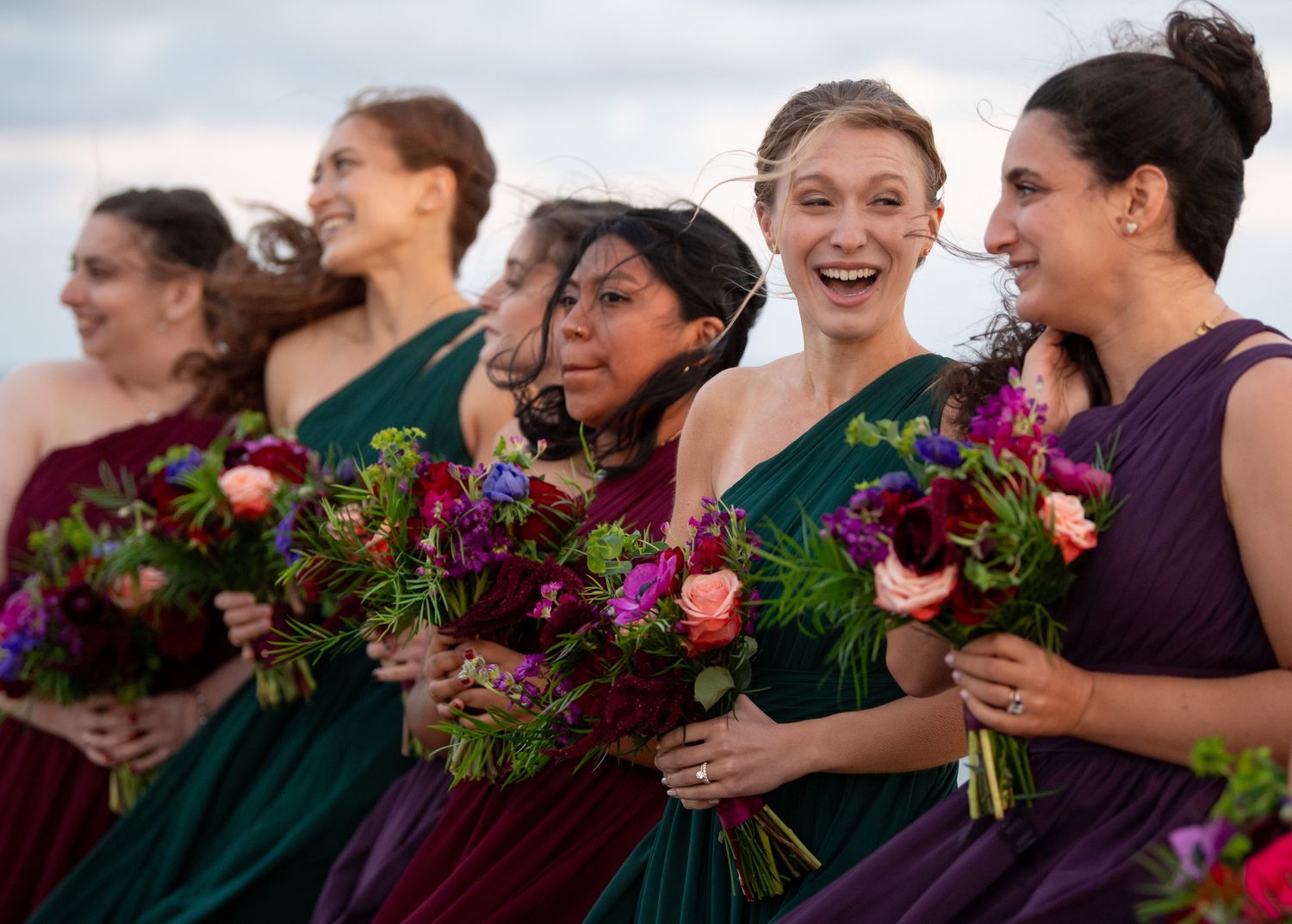 Bridesmaids holding vibrant purple and burgundy floral bouquets at an outdoor wedding.