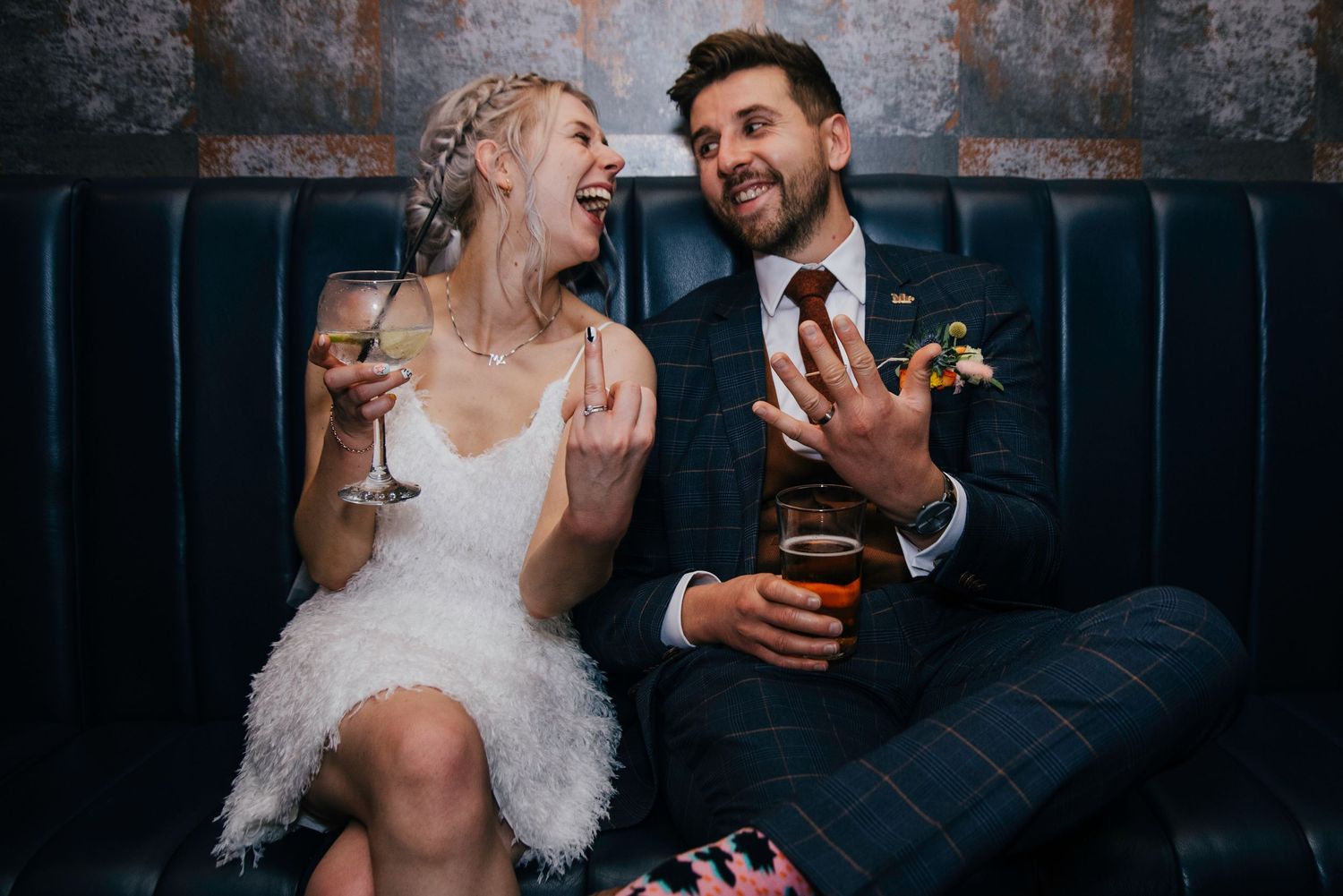 Wedding couple sharing laughs on blue velvet couch during reception.