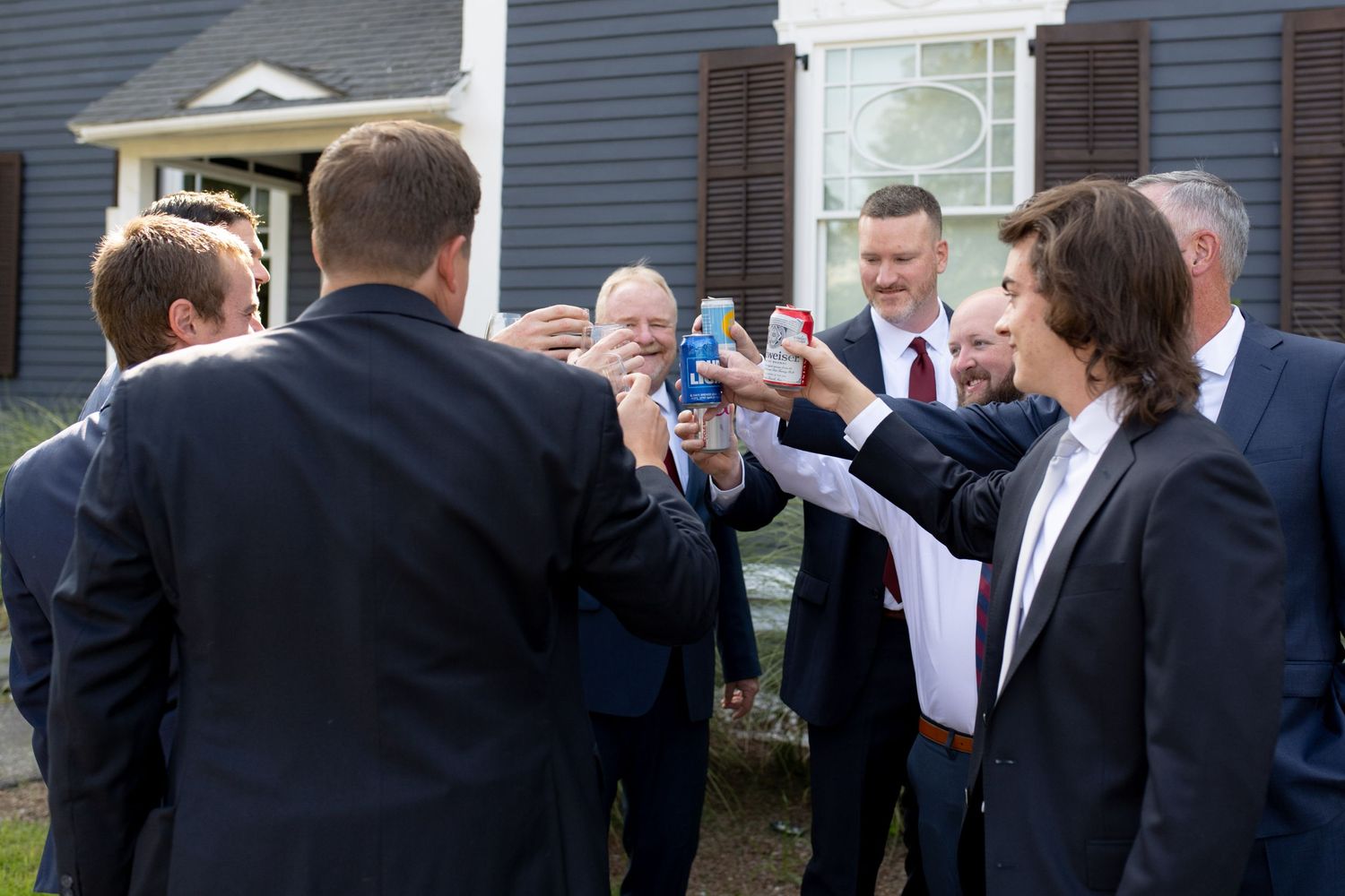 Wedding guests in formal attire gather outside a historic house for celebratory drinks.