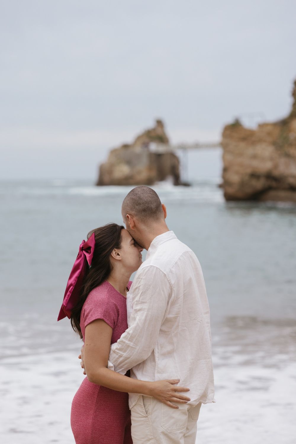 Couple standing together facing the ocean during a soft natural-light engagement session.
