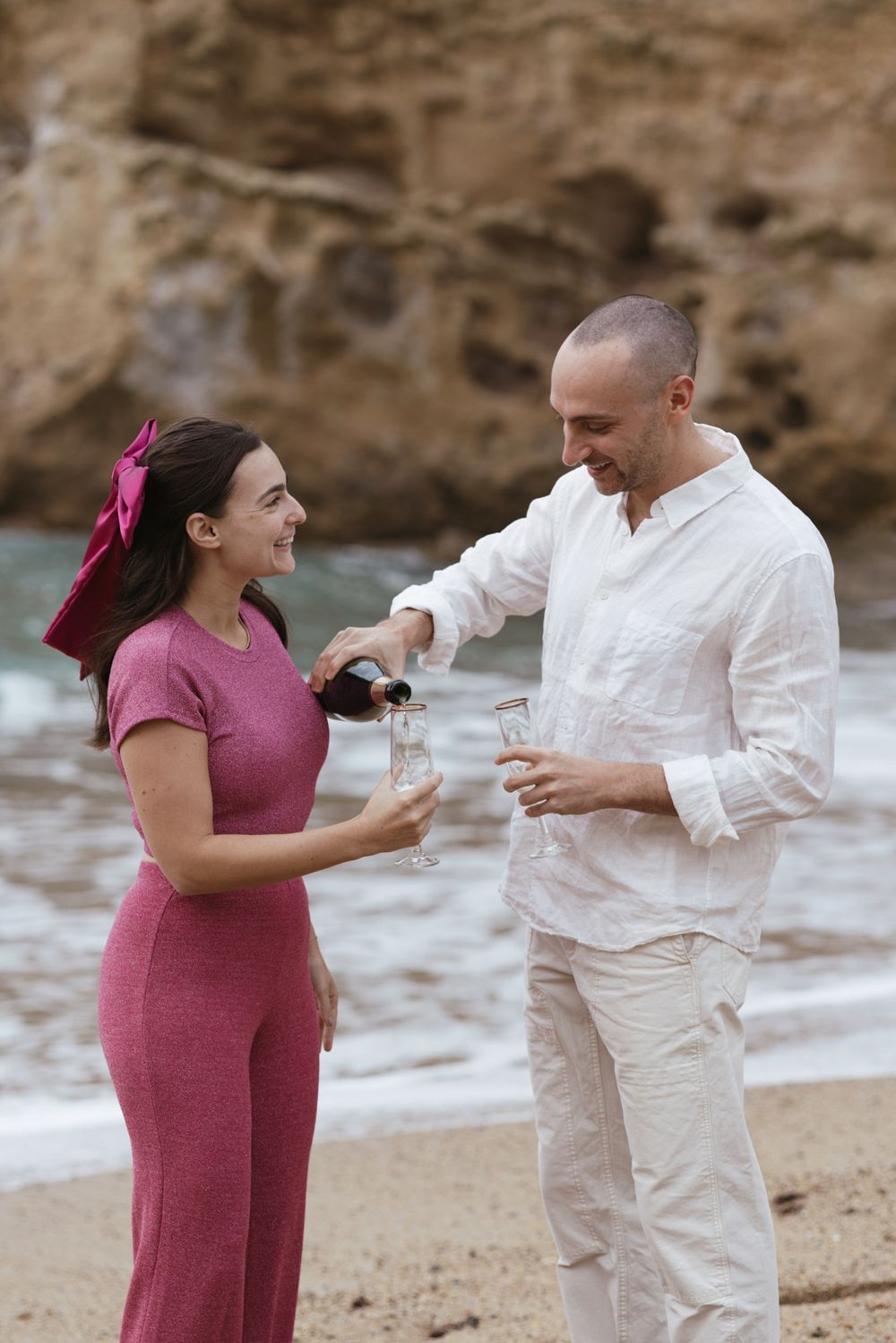 Fine-art engagement photo of a couple opening champagne on a sandy beach, captured with soft natural tones.
