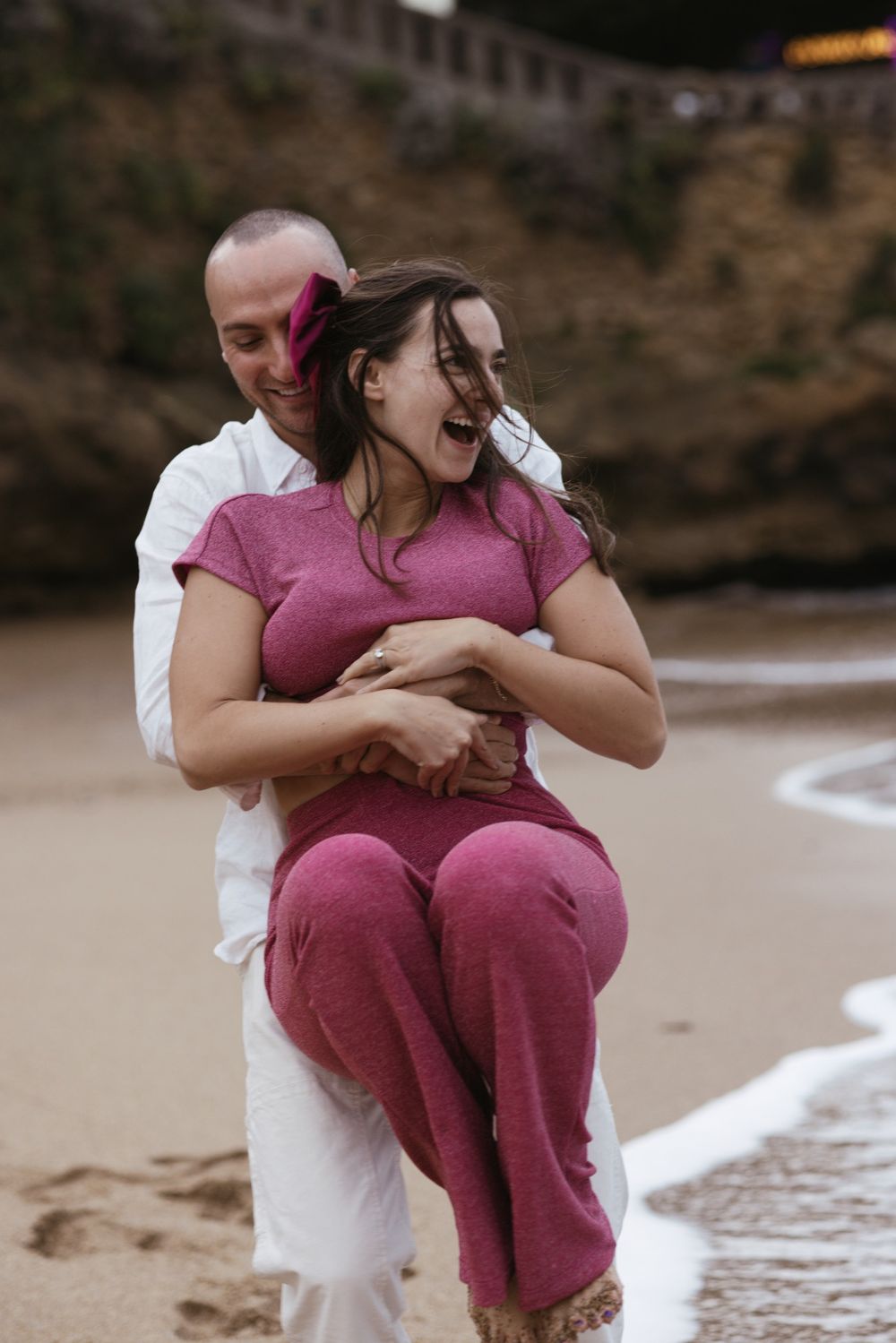 Engagement moment with man lifting his partner playfully on the beach at sunset.