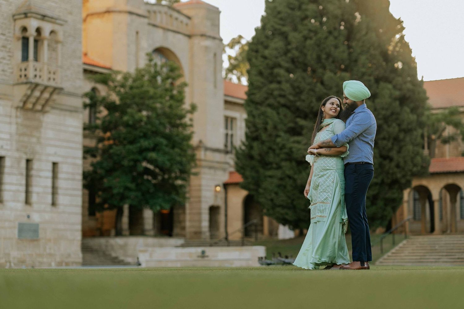 Couple in formal attire embracing on lawn in front of historic castle building.