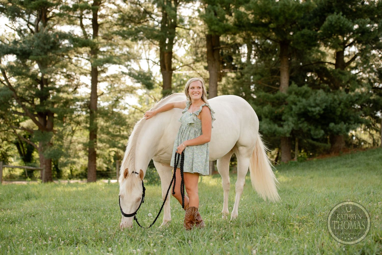 A young woman in a blue dress poses with a white horse in a sunlit forest setting.