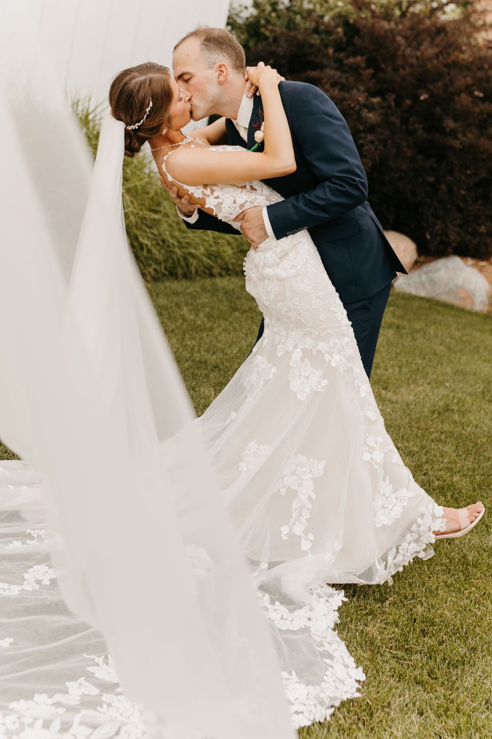 Bride and groom share a romantic dip kiss outdoors, bride wearing lace mermaid gown with flowing veil.
