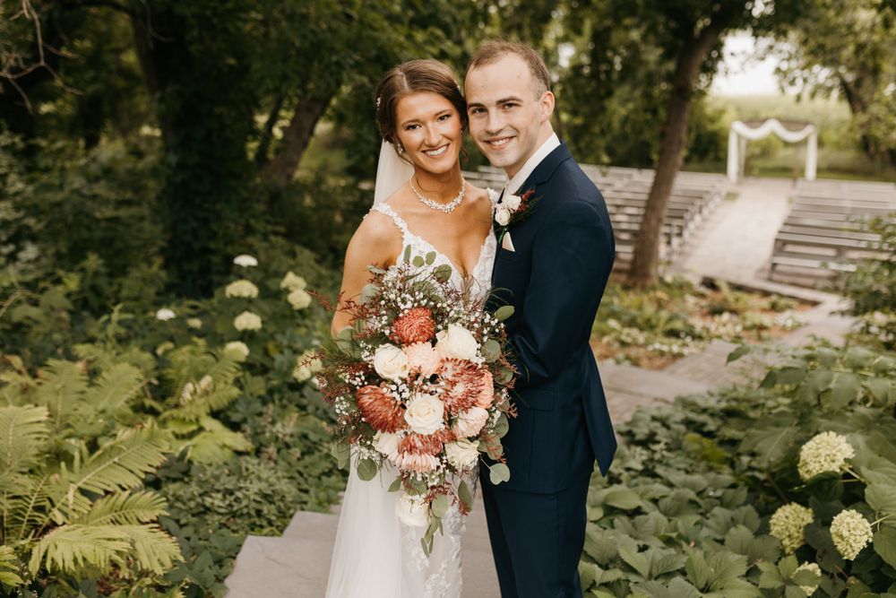 Smiling bride and groom posing together in a garden setting, bride holding a large bouquet of pink and white flowers.