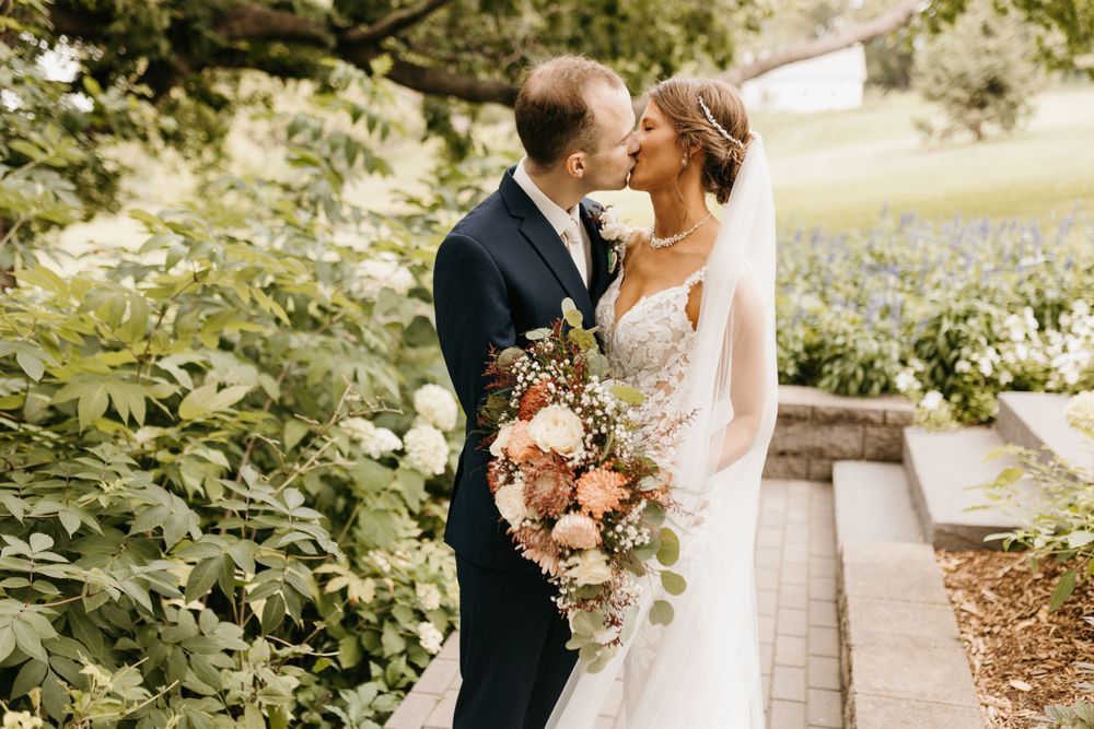 Bride and groom sharing a kiss surrounded by lush greenery, bride holding a beautiful bouquet of pink and rust flowers.