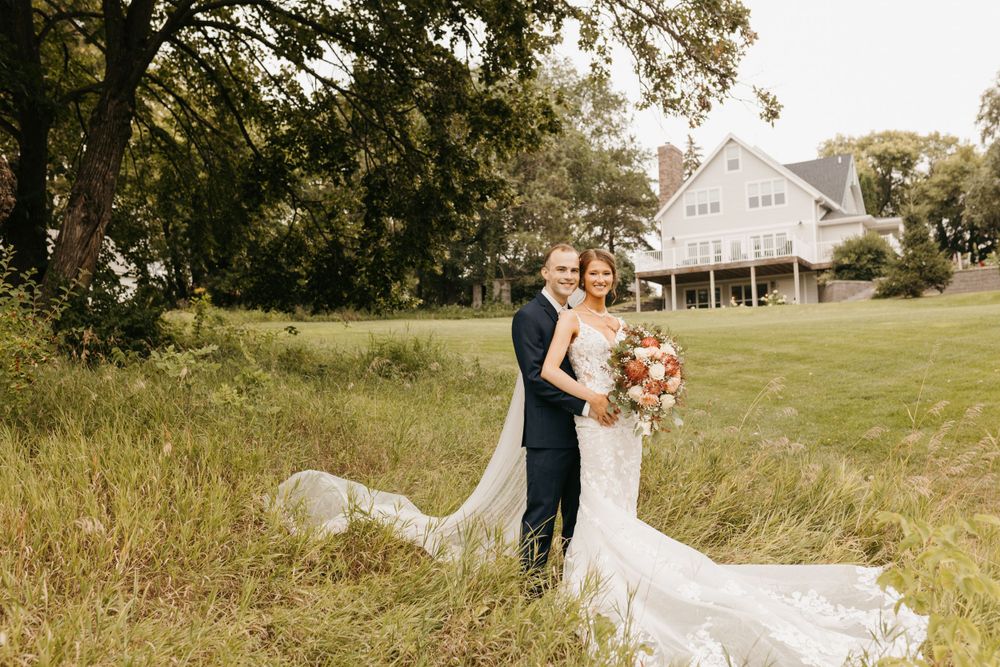 Bride and groom posing in a golden meadow with a white farmhouse in the background, bride holding a floral bouquet.