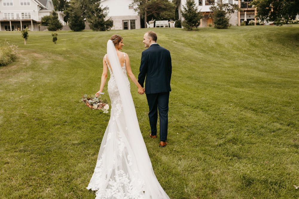 Bride in lace gown with cathedral veil holds bouquet while walking with groom on lawn.