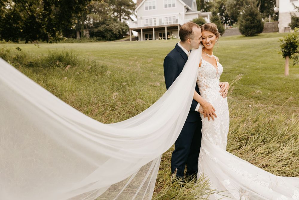 Bride in lace gown with flowing veil embraces groom in navy suit on green meadow.