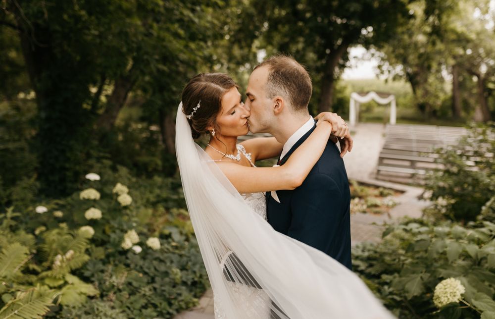 Bride and groom sharing a kiss in a lush garden, bride's veil flowing gracefully with an outdoor altar in background.