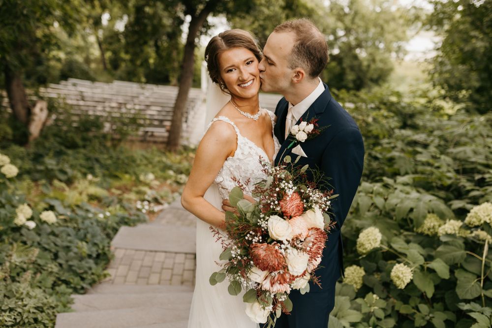 Groom kissing bride on the cheek in a lush garden, bride smiling and holding a bouquet of pink and peach flowers.