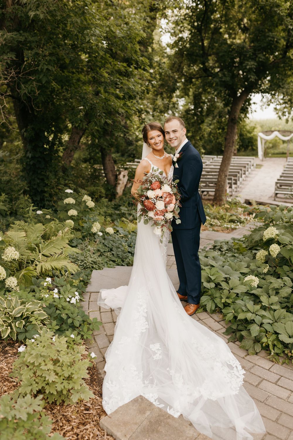 Bride and groom standing on a garden path, bride in a lace gown with a long train, holding a colorful bouquet.