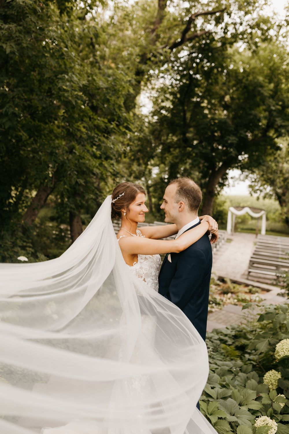 Bride and groom facing each other romantically with the bride's veil flowing in the wind in a garden setting.