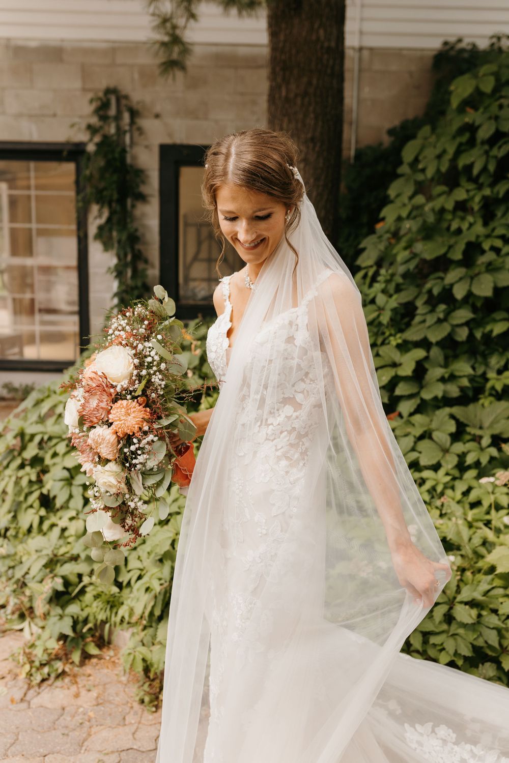 Smiling bride in lace gown holds colorful floral bouquet with flowing veil outdoors.