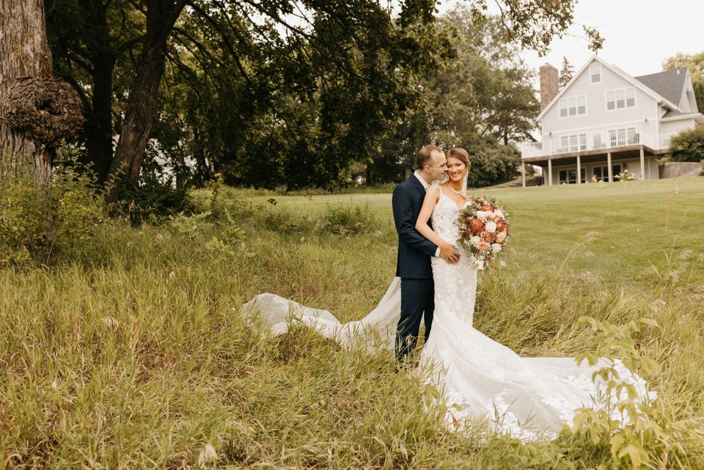Bride and groom standing in a sunlit grassy field near a white farmhouse, bride's lace train flowing behind her.