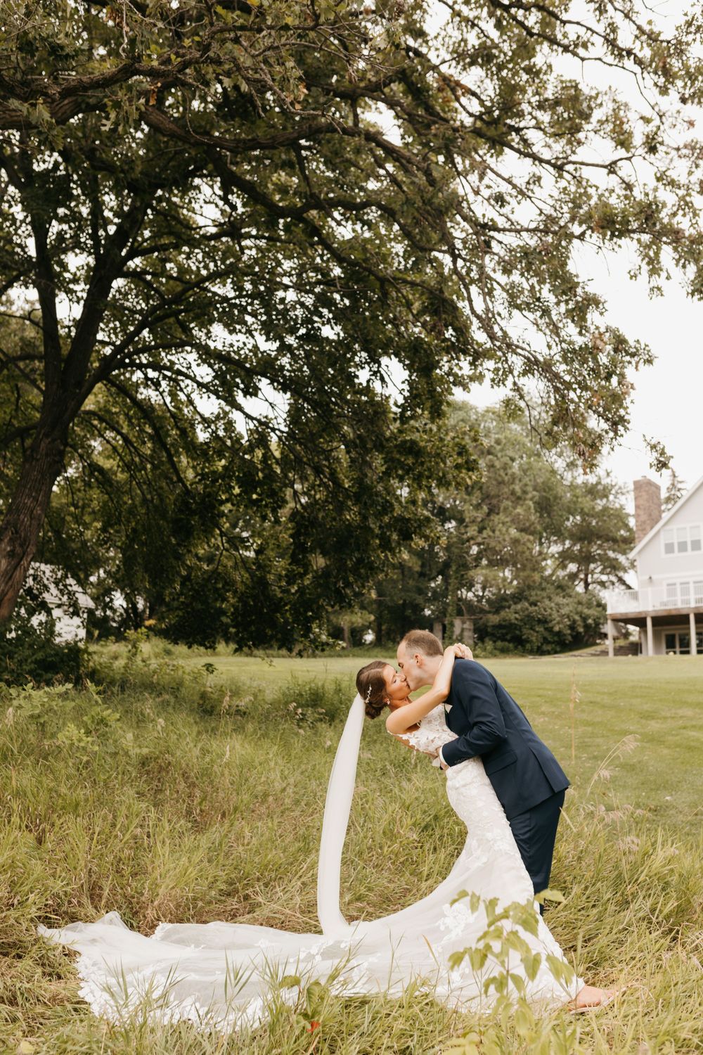 Groom dips bride for romantic kiss in lace wedding gown with long veil outdoors.