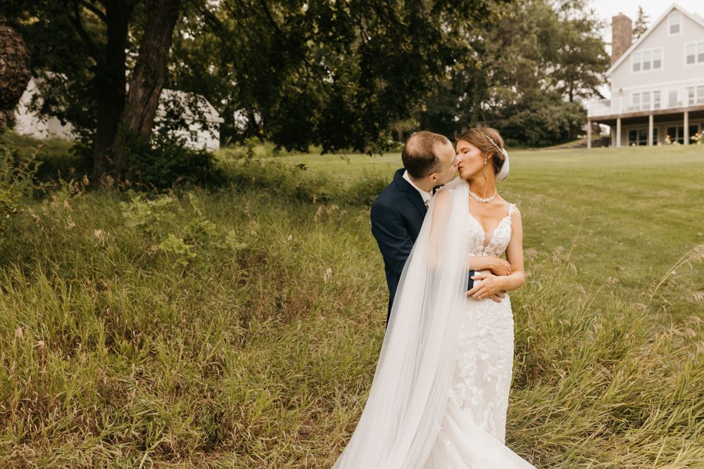 Groom kisses bride on cheek as she smiles in white gown with veil in grassy field.