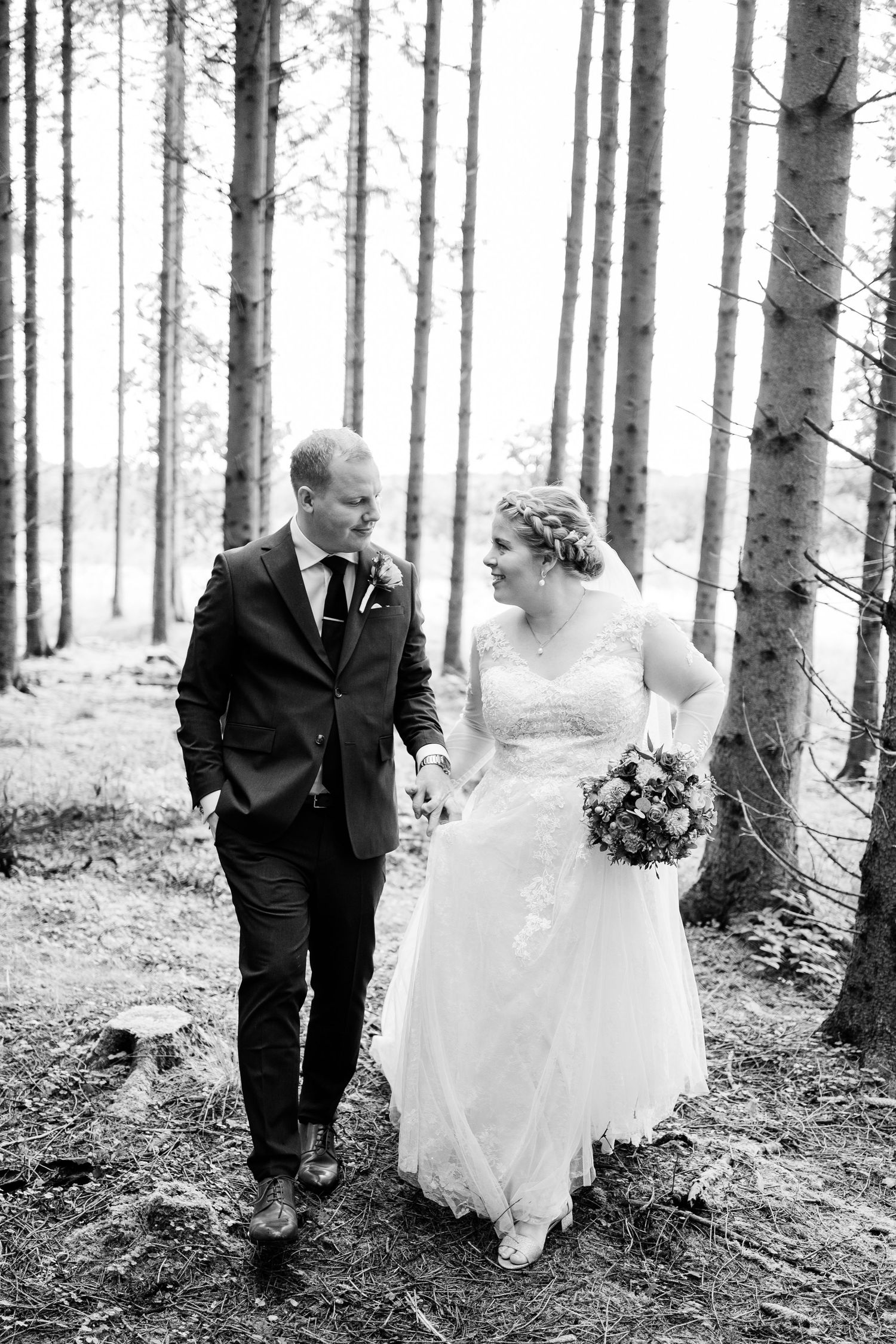 A romantic black and white photo of newlyweds walking together through a serene forest setting.